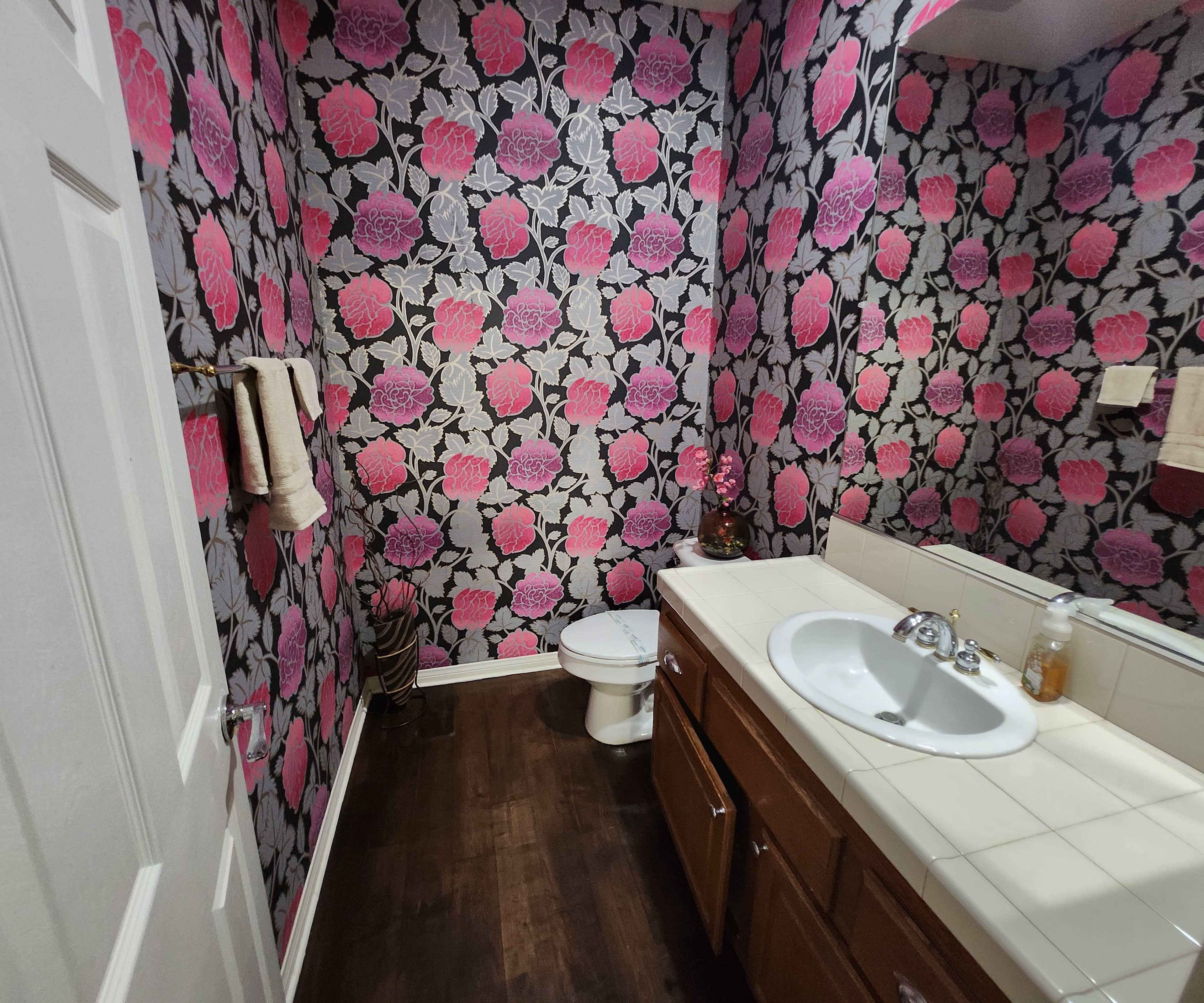 A small bathroom with floral wallpaper featuring large pink roses, a white sink with a mirror above it, and a toilet.
