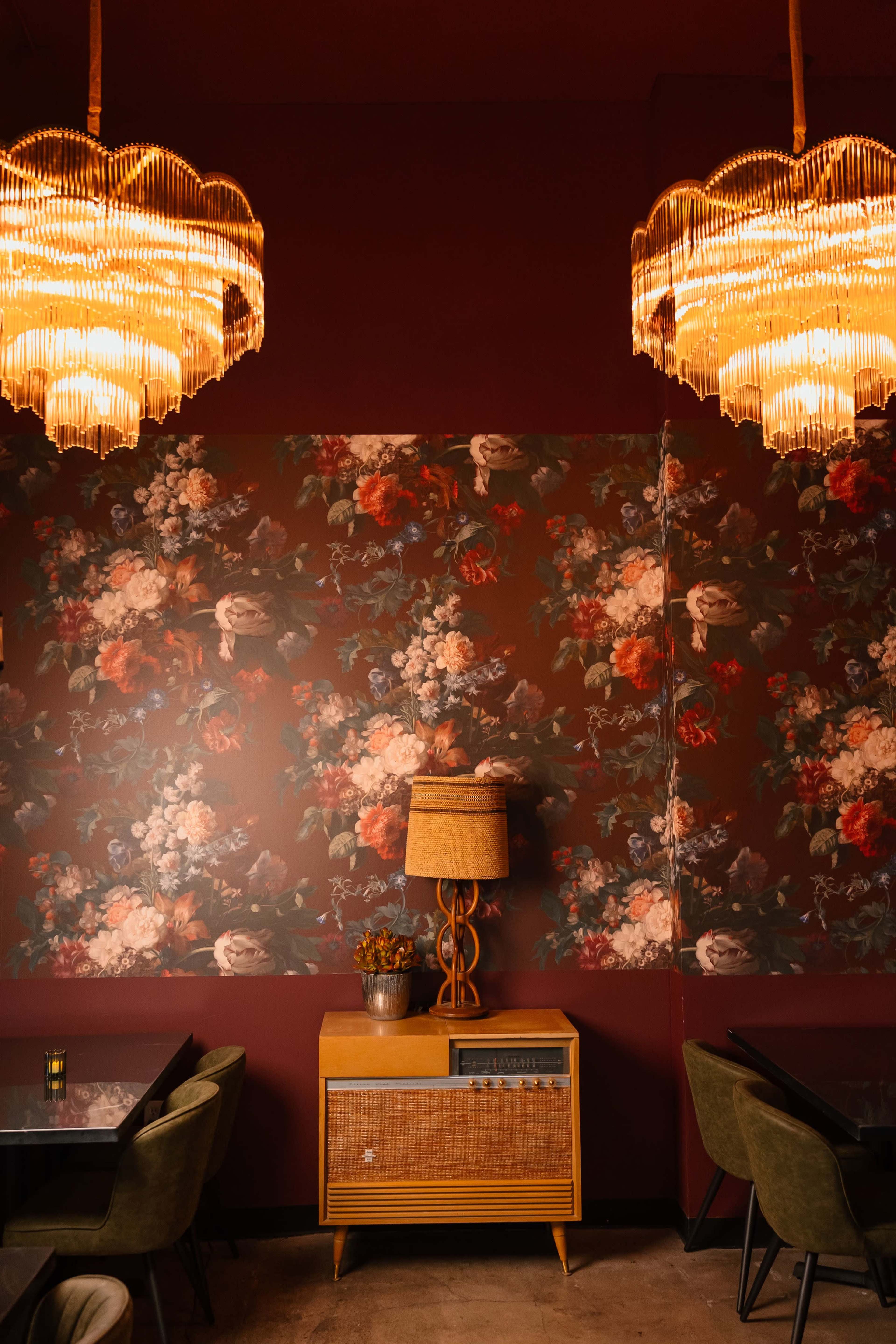 The image shows a cozy restaurant interior featuring a floral wallpaper, a vintage lamp, and two ornate chandeliers.