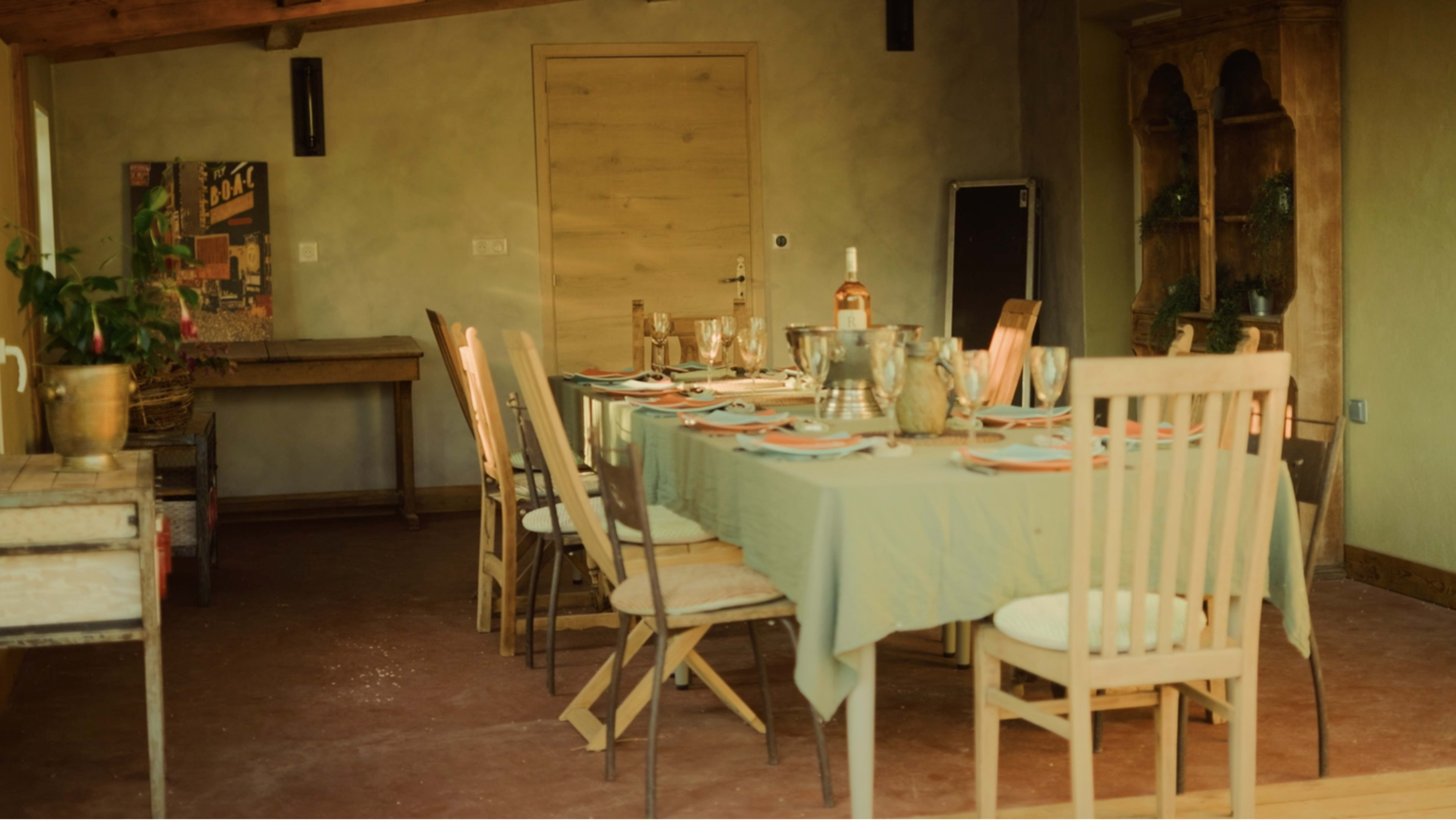 A long dining table is set in a softly lit room, with chairs arranged around it, and various dinnerware and glasses placed on the table.