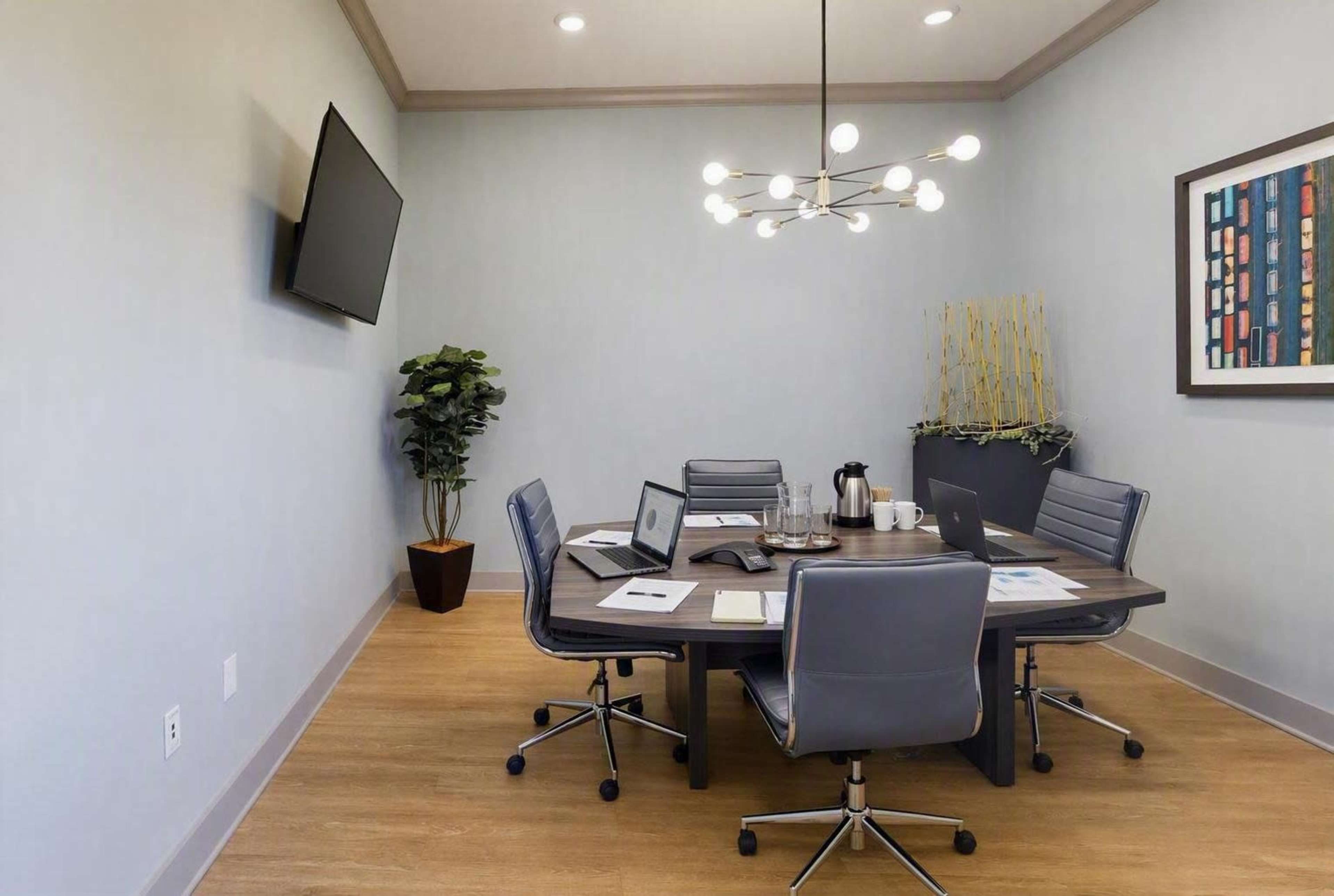 A modern conference room features a round table surrounded by four chairs, with a large television mounted on the wall and decorative plants in the corner.