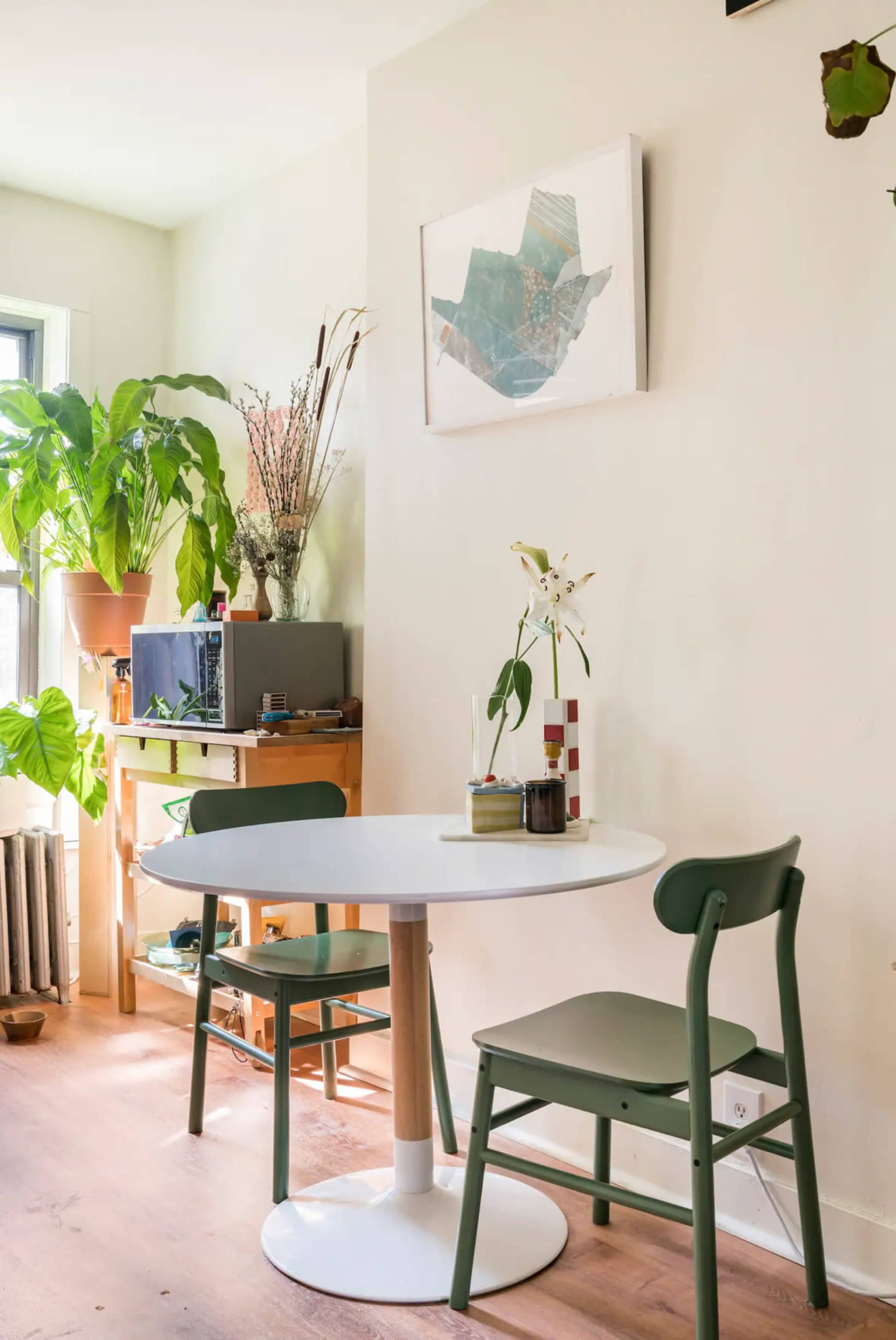 A round white table with two green chairs is positioned near a wall adorned with a piece of art, alongside various indoor plants and a shelf with decorative items.