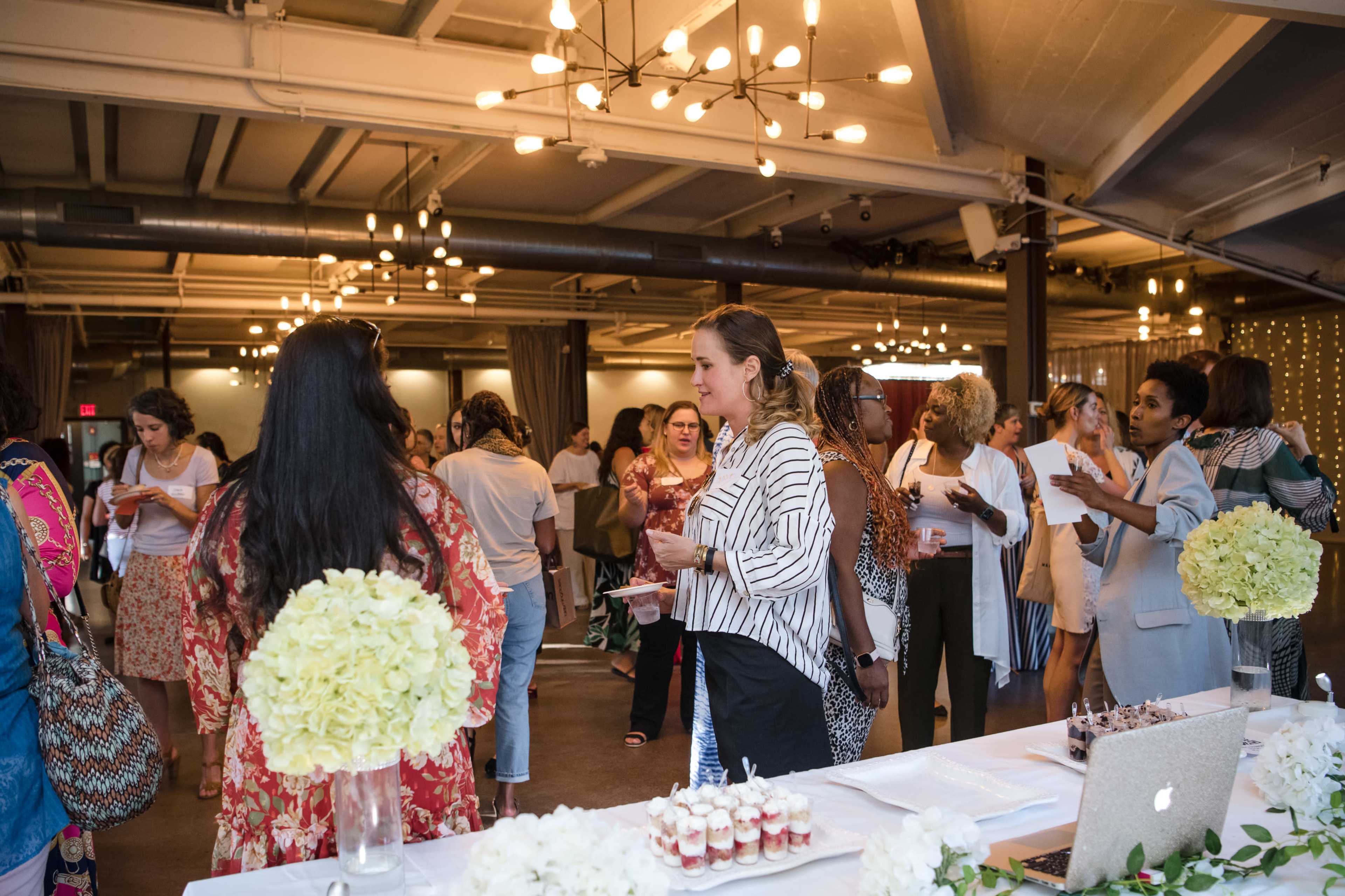 A group of people mingles at a well-lit event space featuring decorated tables with flowers and refreshments.
