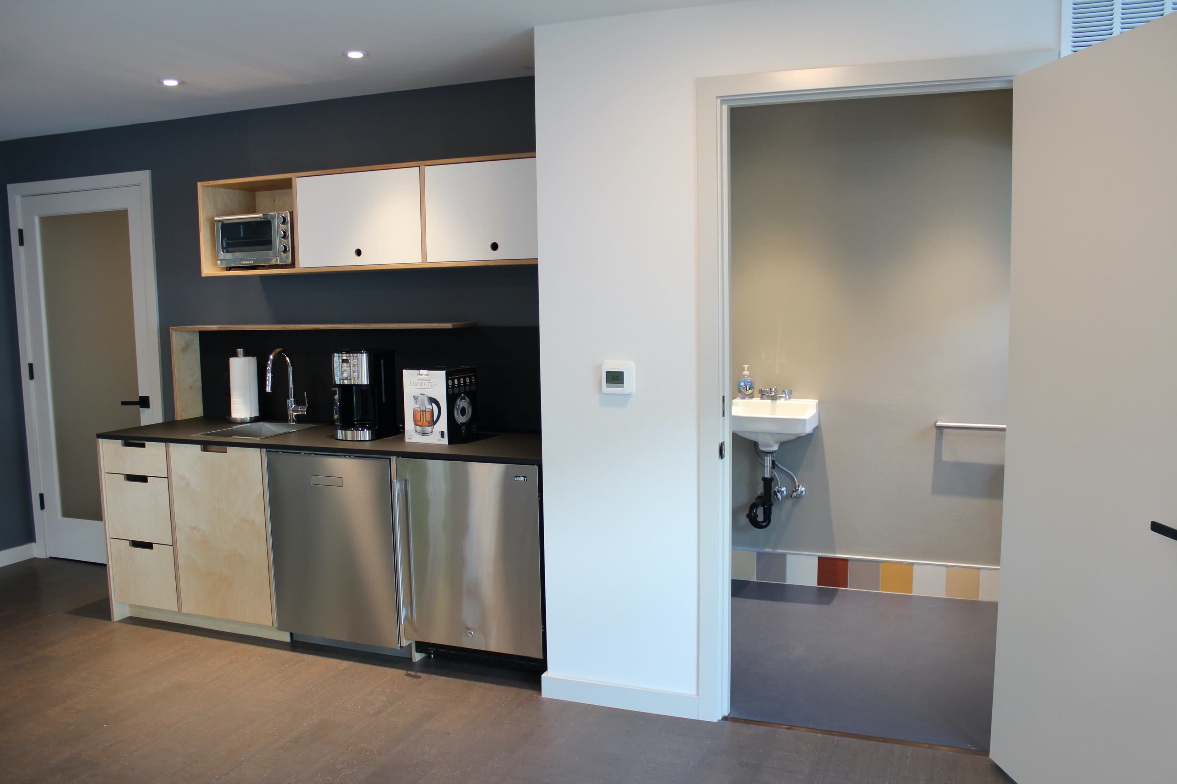 The image shows a modern kitchen area with a stainless steel refrigerator, microwave, and a small sink, adjacent to a bathroom with a toilet and grab bars.