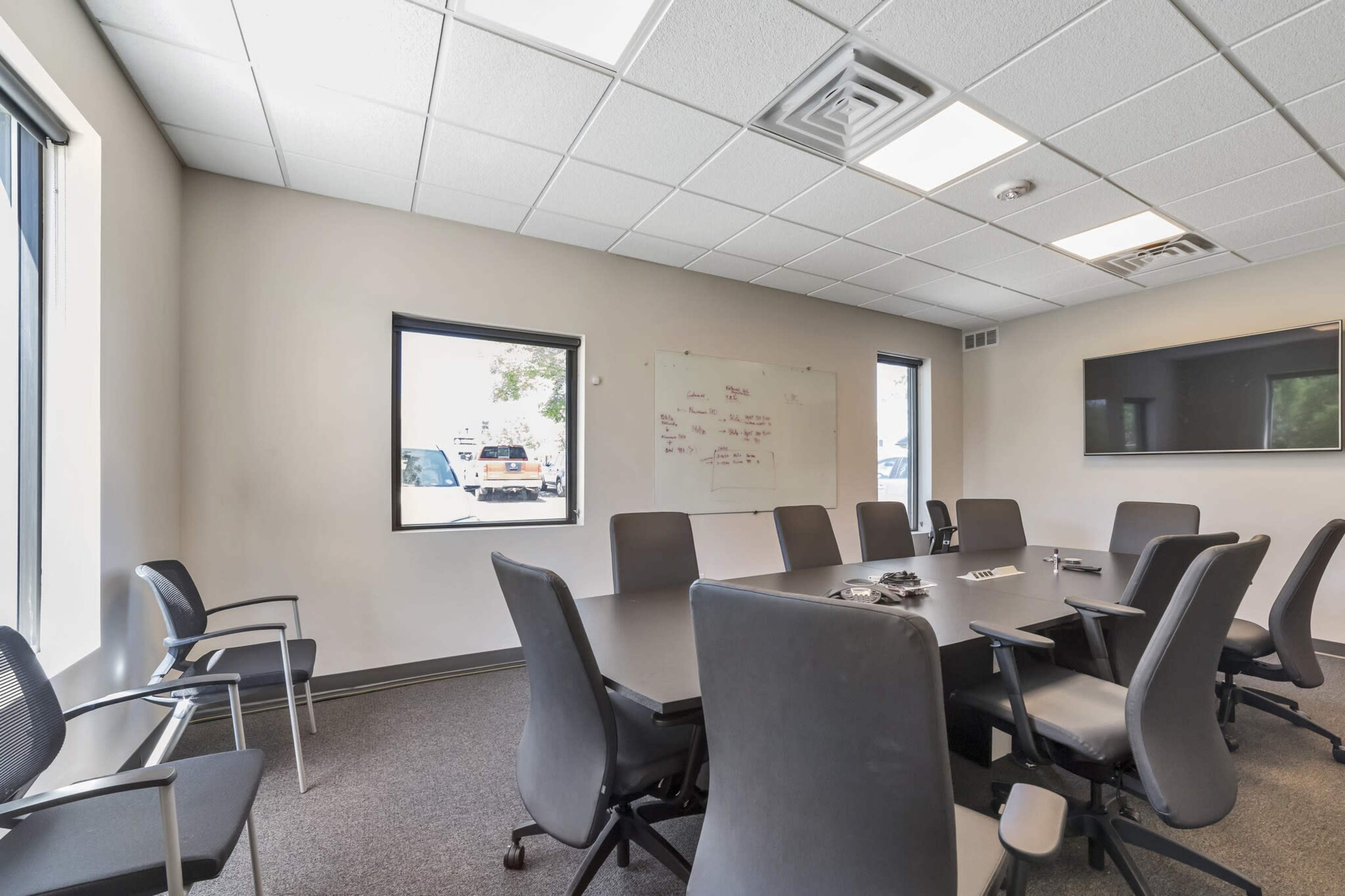 The image shows a modern conference room with a large table, several chairs, a whiteboard, and a wall-mounted screen.
