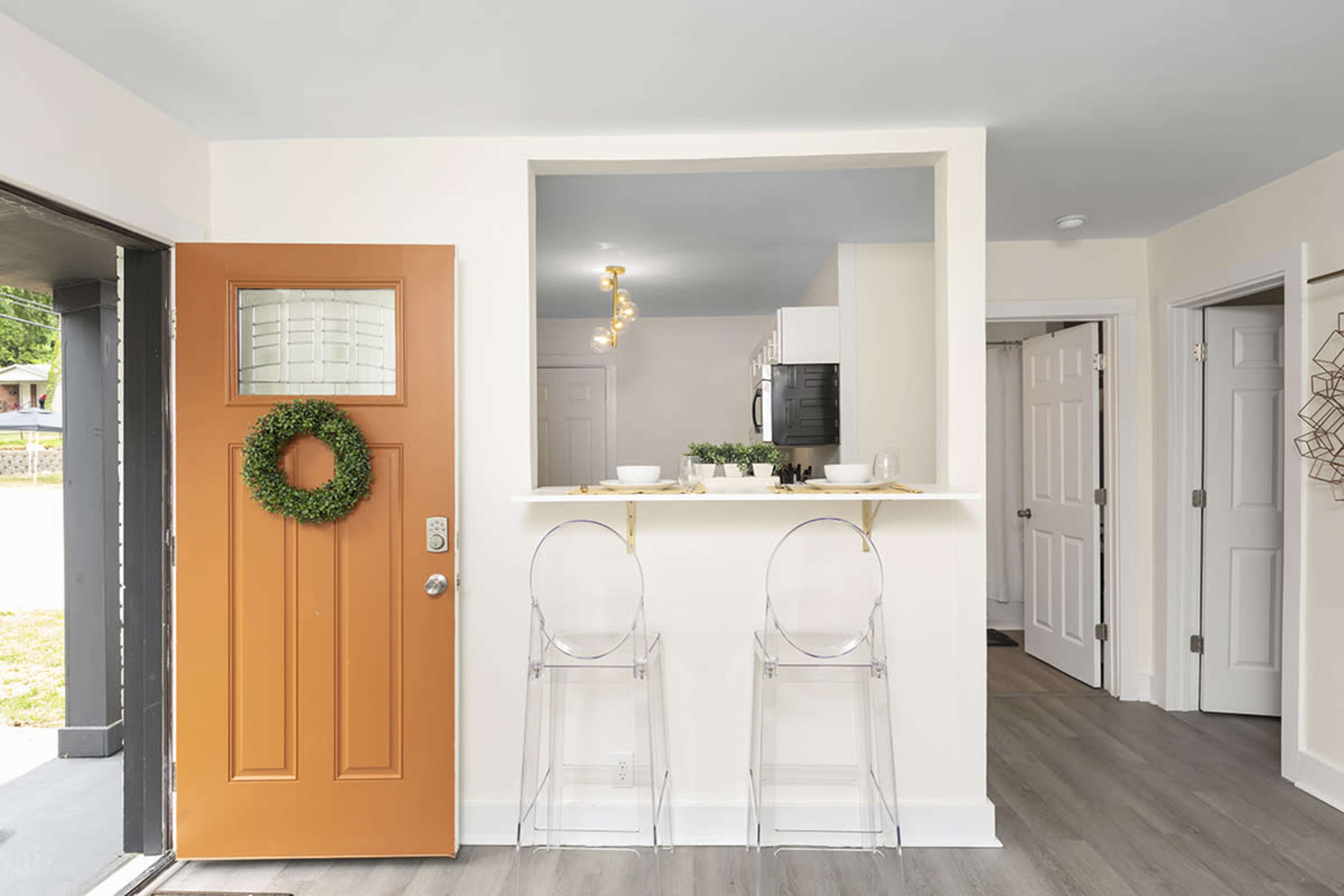 An entryway with an orange front door, a small bar area with two clear chairs, and an adjoining kitchen space in a bright room.