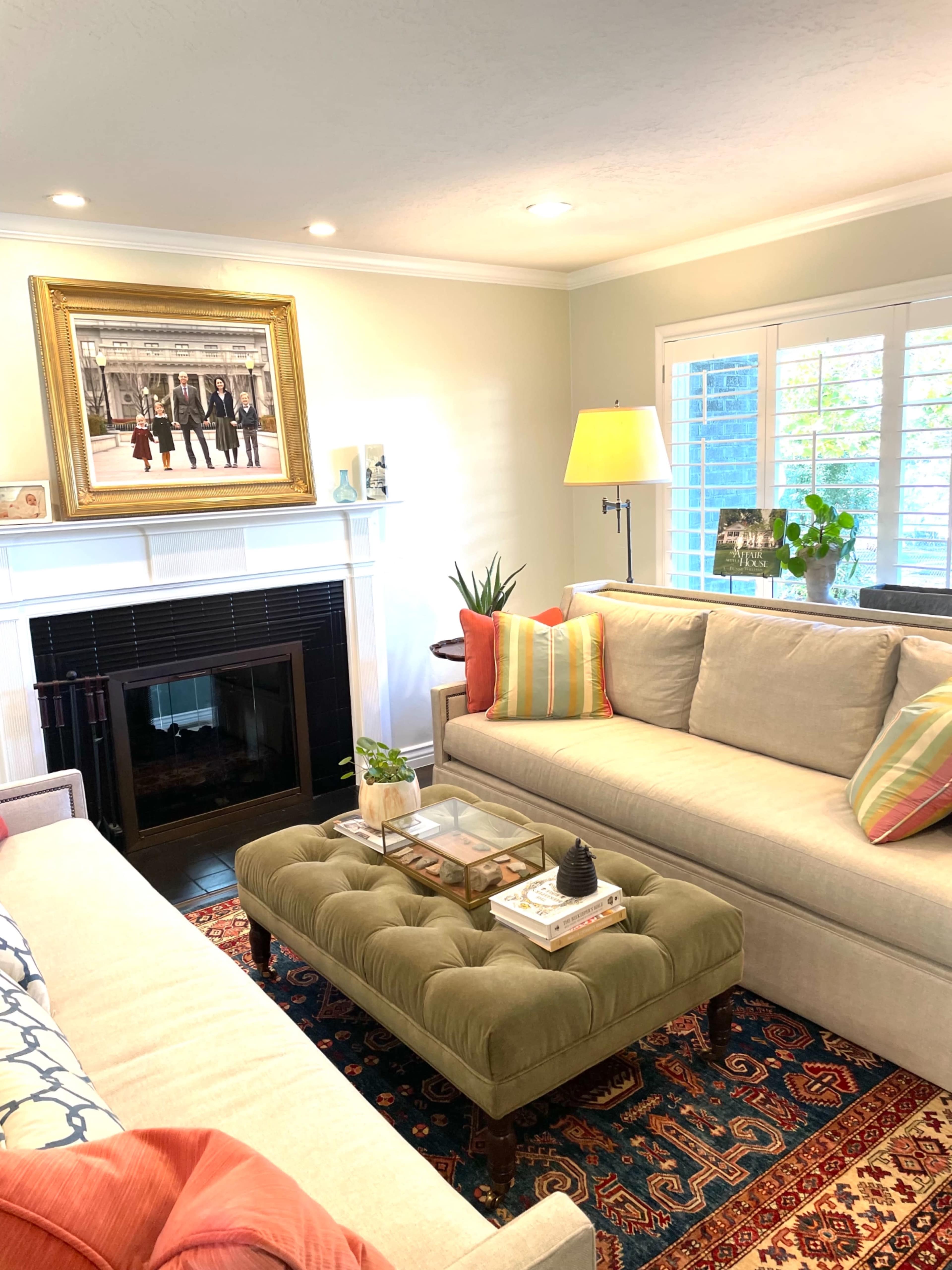 A bright living room features two sofas, a tufted coffee table, a framed photo above a fireplace, and natural light coming through window shutters.
