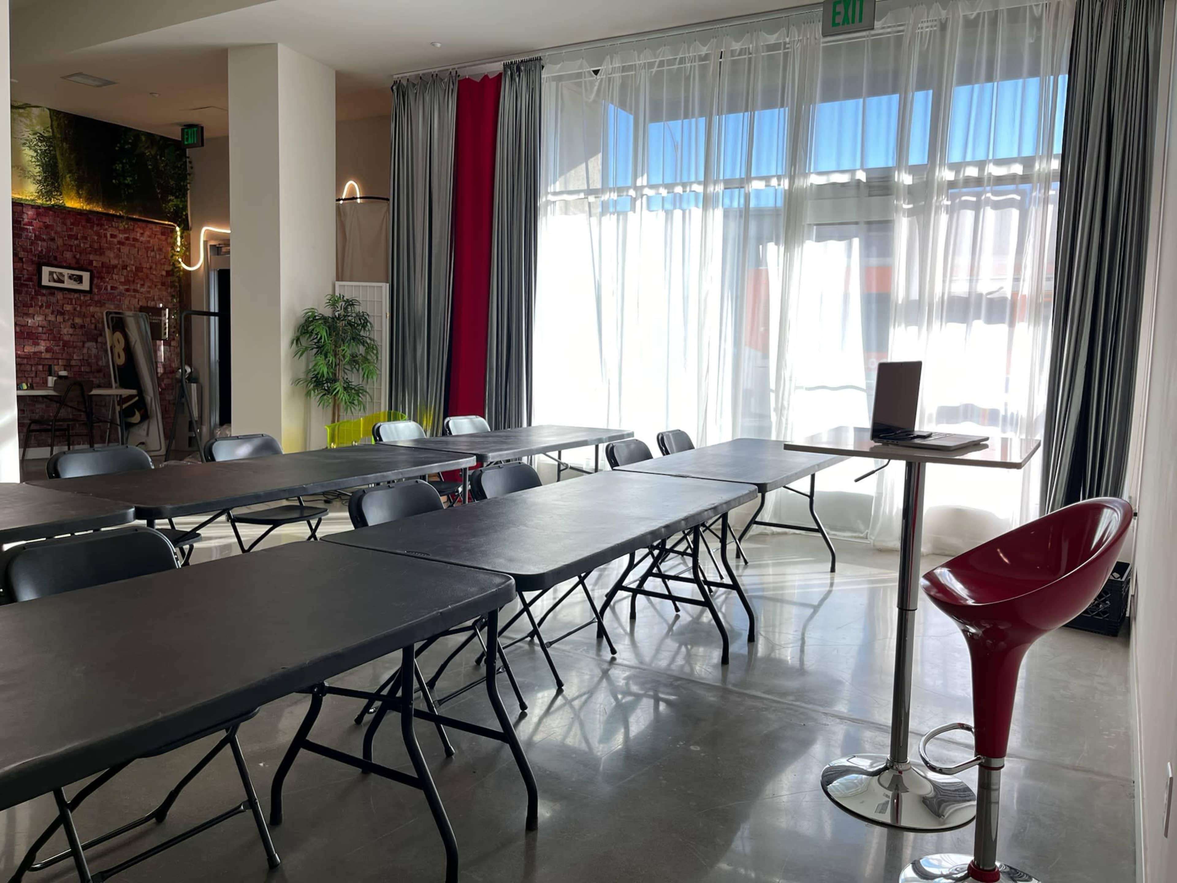 A bright meeting room with several foldable tables arranged in a row and a tall bar stool beside a laptop on a small table.