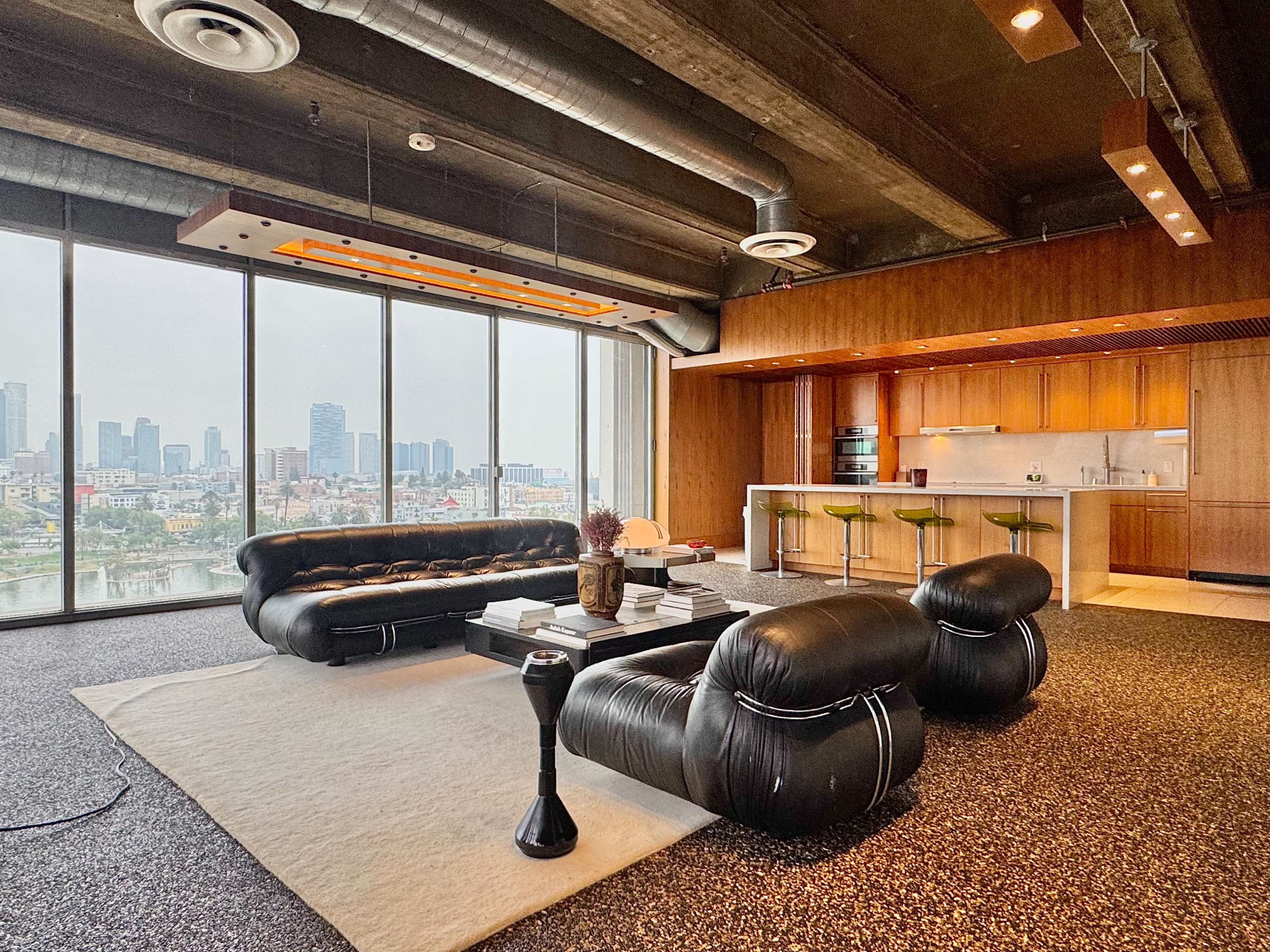 The image shows a modern living room with large windows overlooking a city skyline, featuring a dark, curved sofa, a white area rug, and a kitchen area with wooden cabinets and bar seating.
