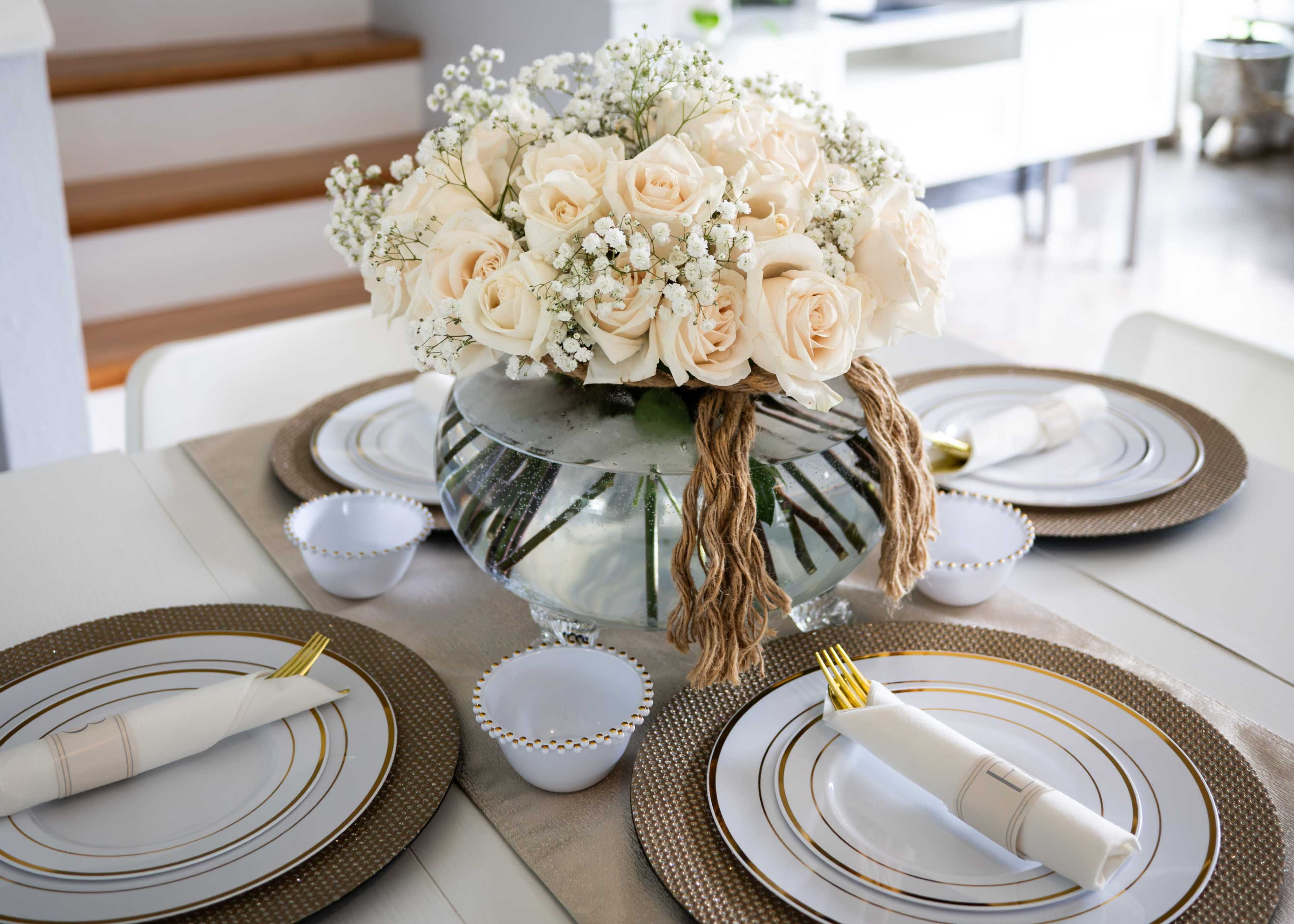 A dining table is set with elegant dishes and a large floral arrangement of white roses in a glass vase.