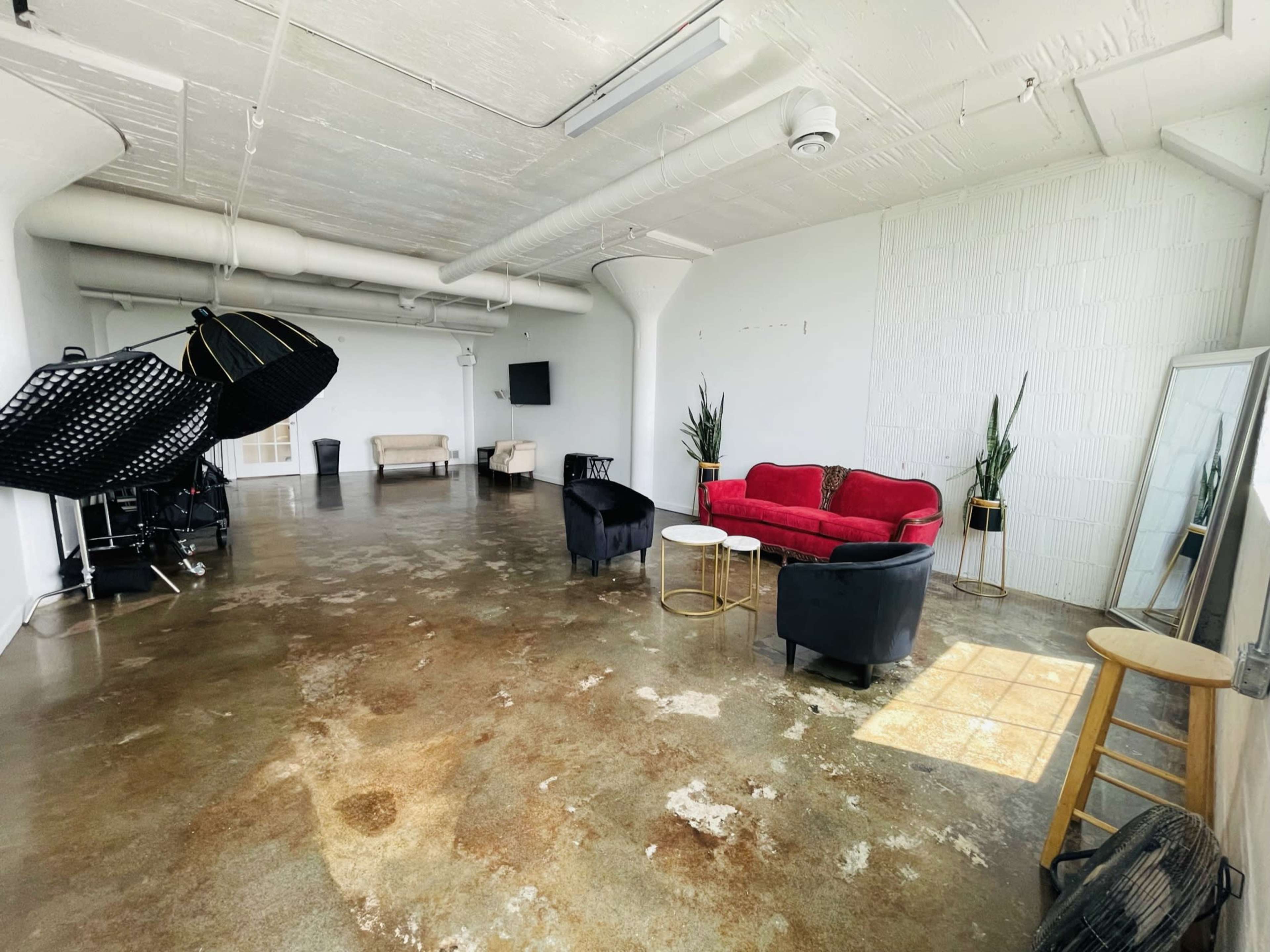 An empty studio space with a polished concrete floor, two black chairs, a red sofa, small tables, a large mirror, and lighting equipment.