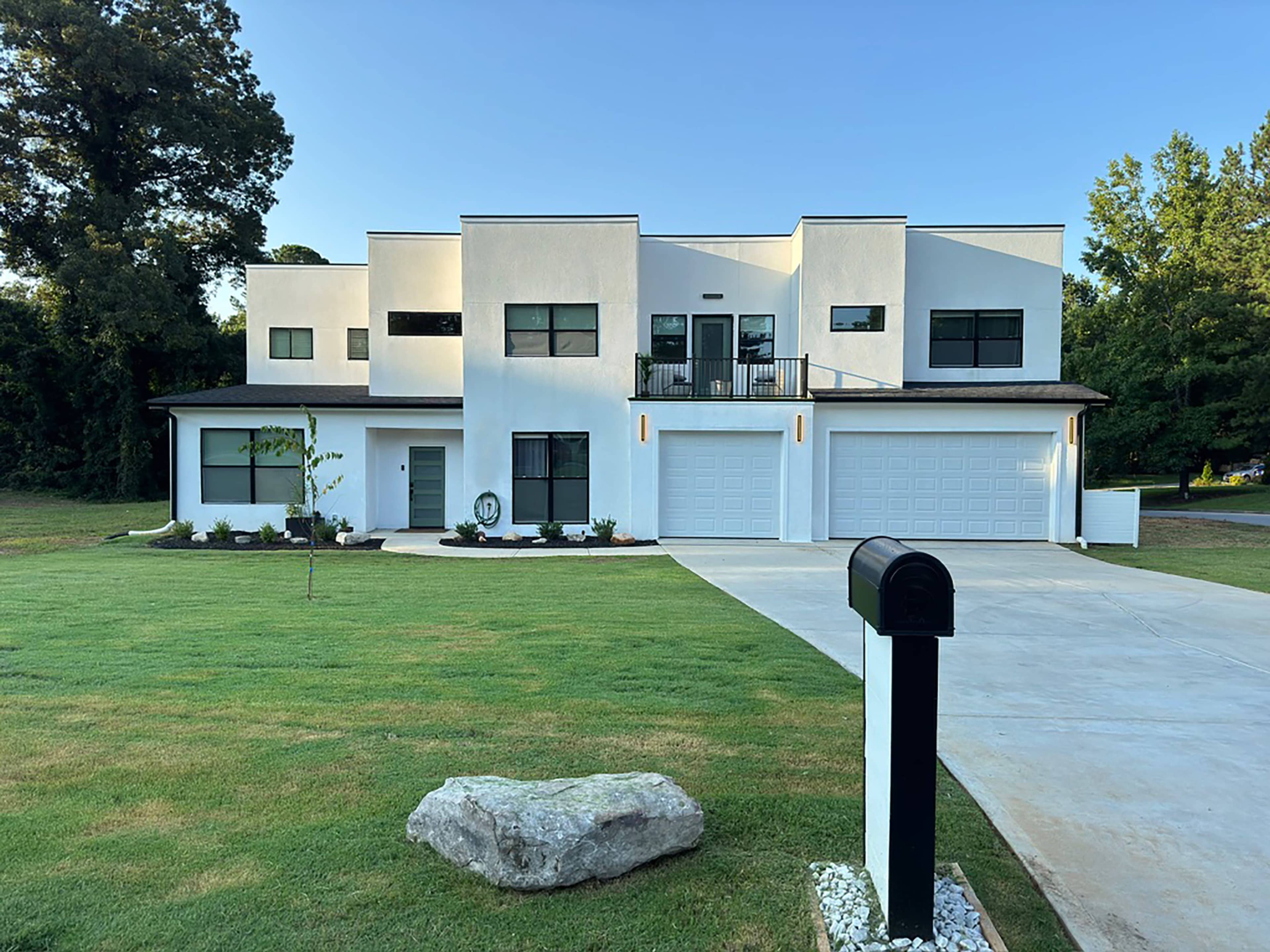 A modern two-story house with a white exterior features large windows, a balcony, and a three-car garage on a green lawn.