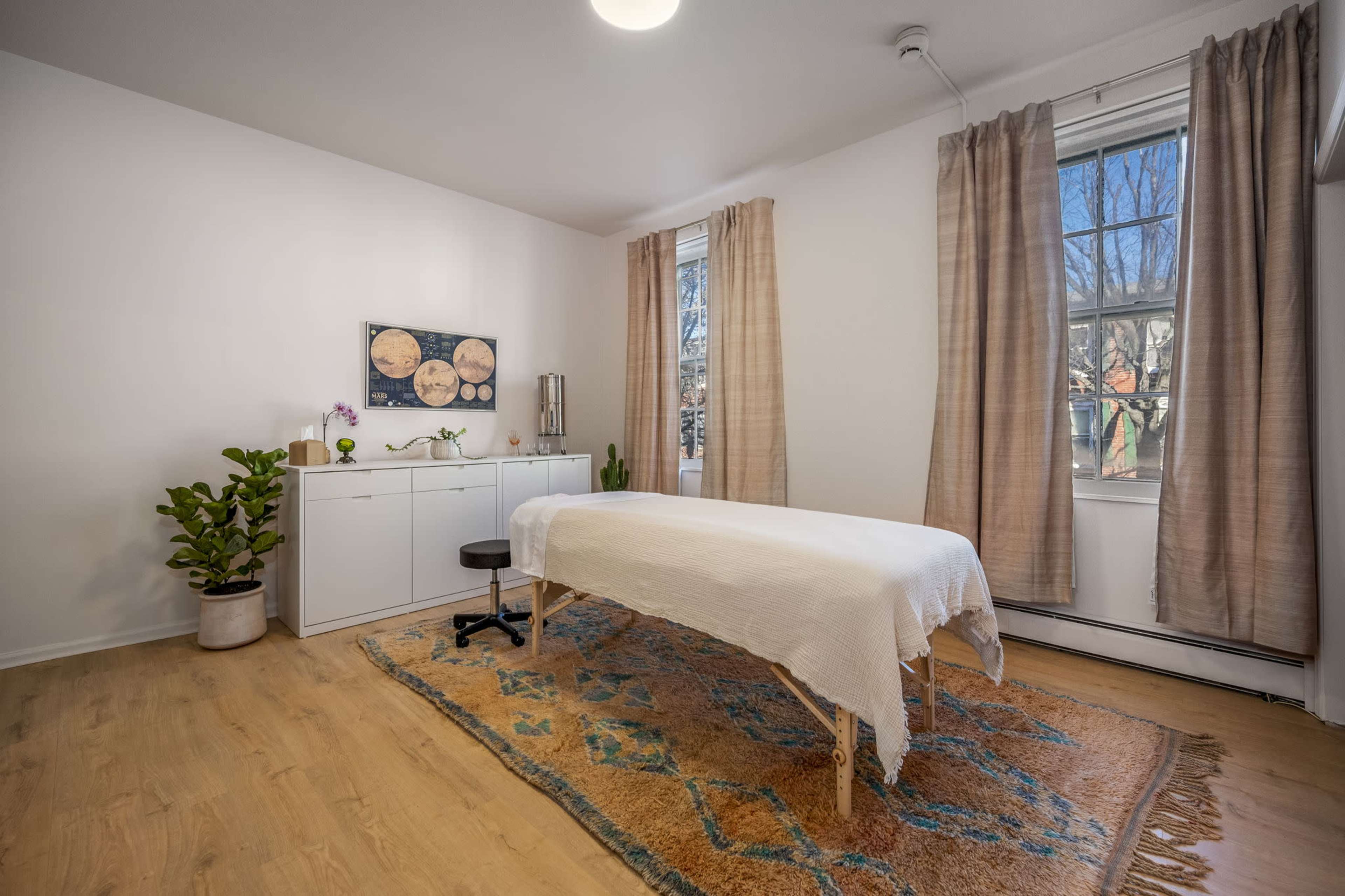 The image shows a light-colored room with a massage table covered by a white sheet, surrounded by a rug, plants, and a window with natural light.