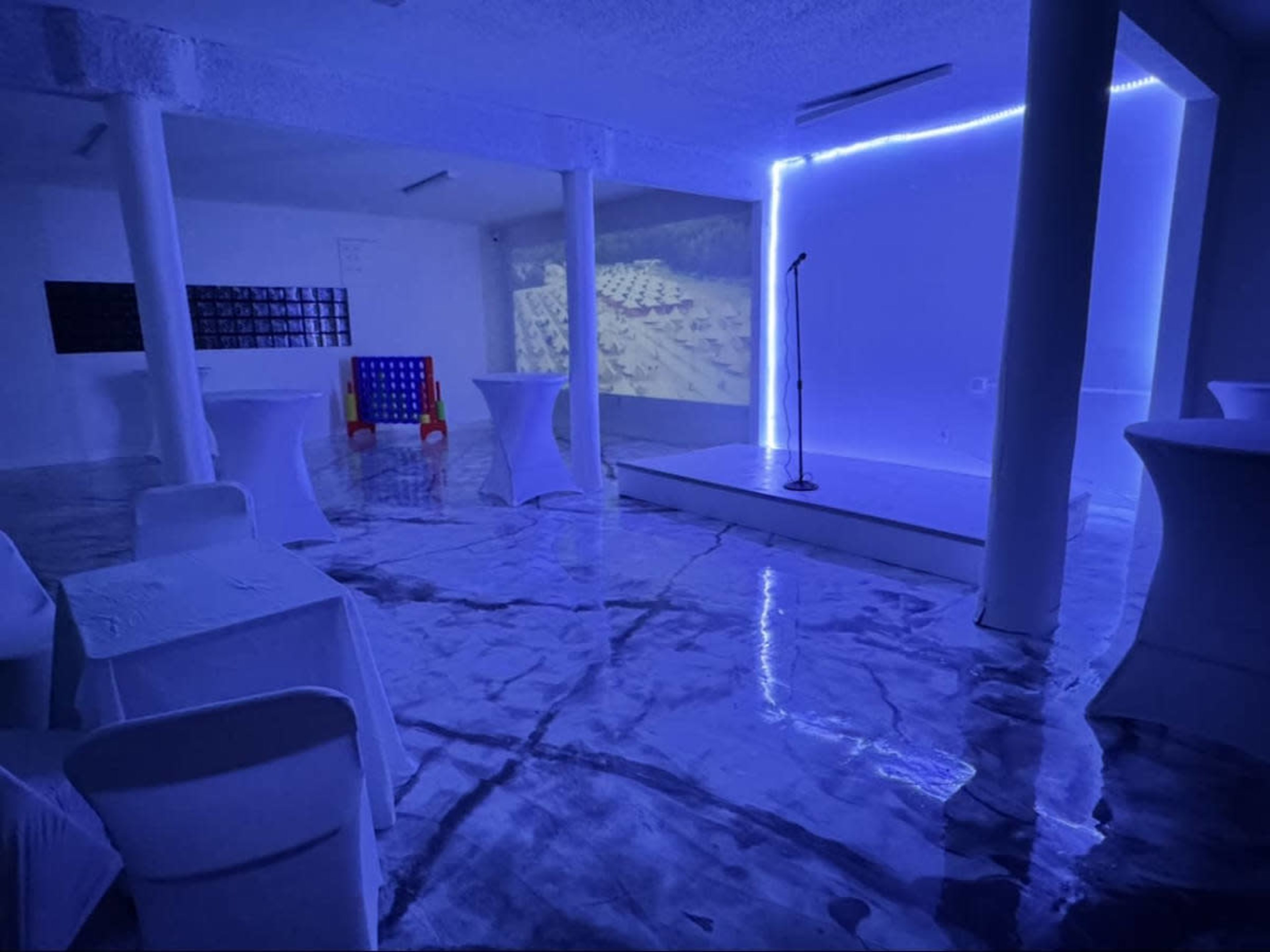 The image shows a modern, brightly lit room with blue lighting, featuring a small stage with a microphone, several white tables, and a projected screen on the wall.