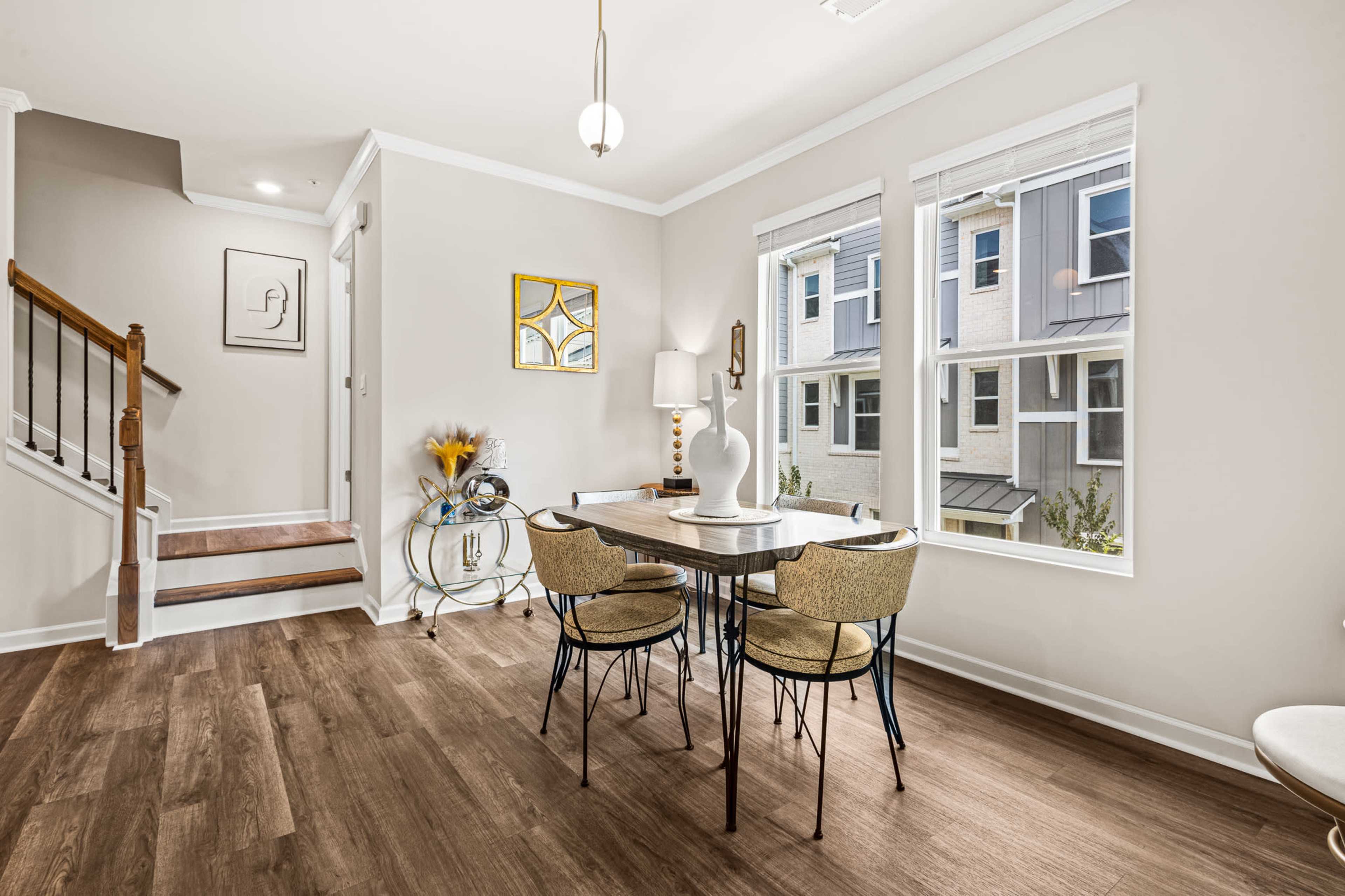 A dining area features a circular table with four chairs, situated near windows that provide natural light, alongside a staircase and decorative elements on the walls.