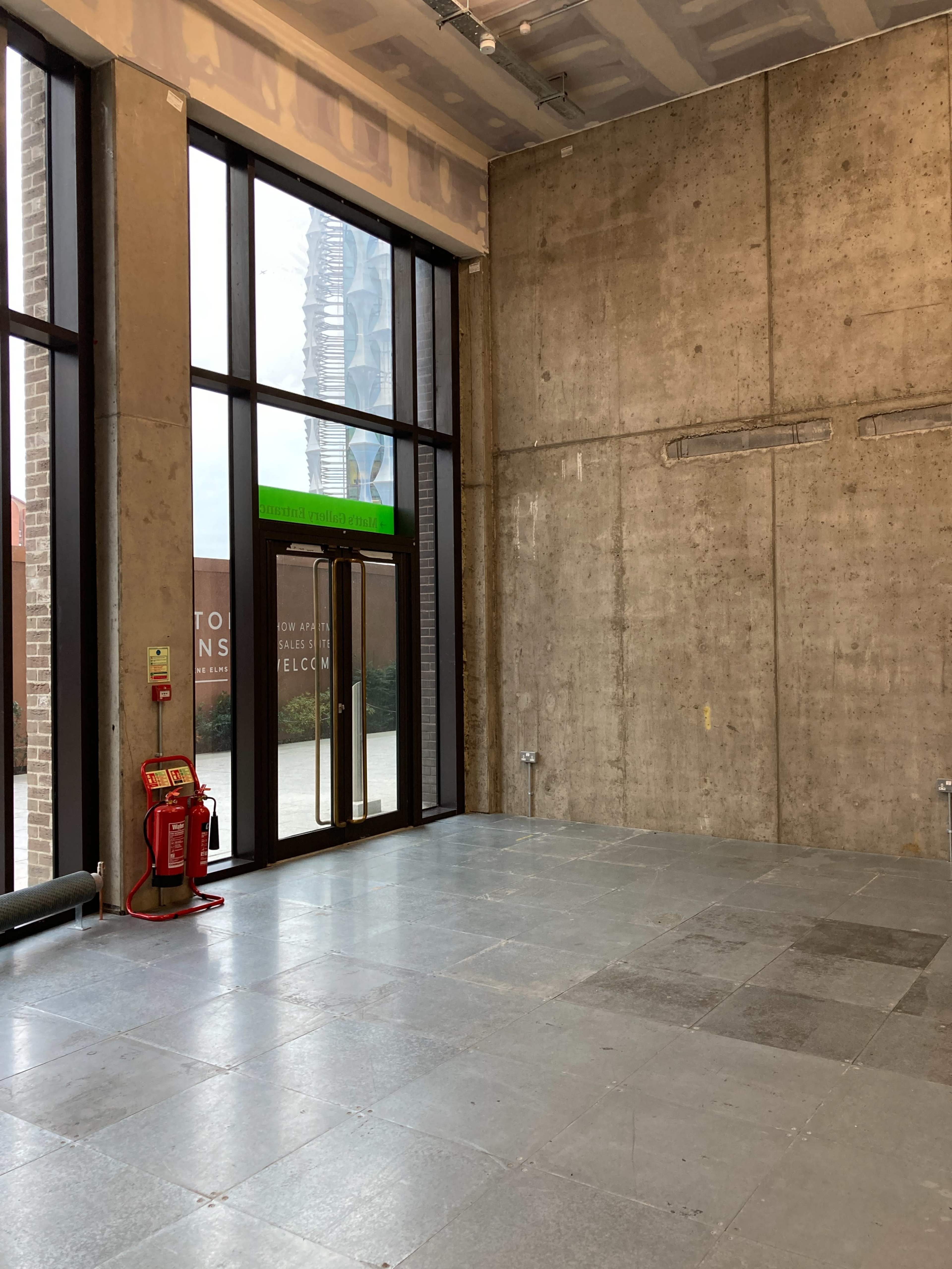 An empty room with concrete walls and large windows revealing an outdoor view, featuring a fire extinguisher near the entrance.