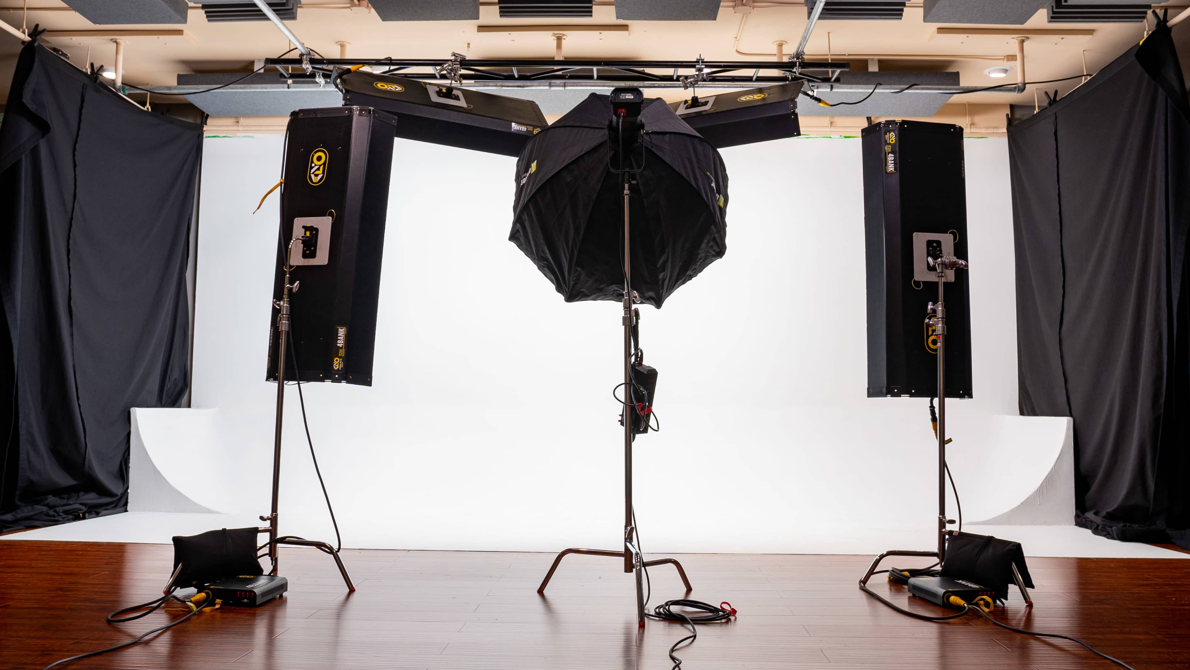 A photography studio setup featuring three lighting equipment stands and a large softbox on a central tripod, all facing a white backdrop.