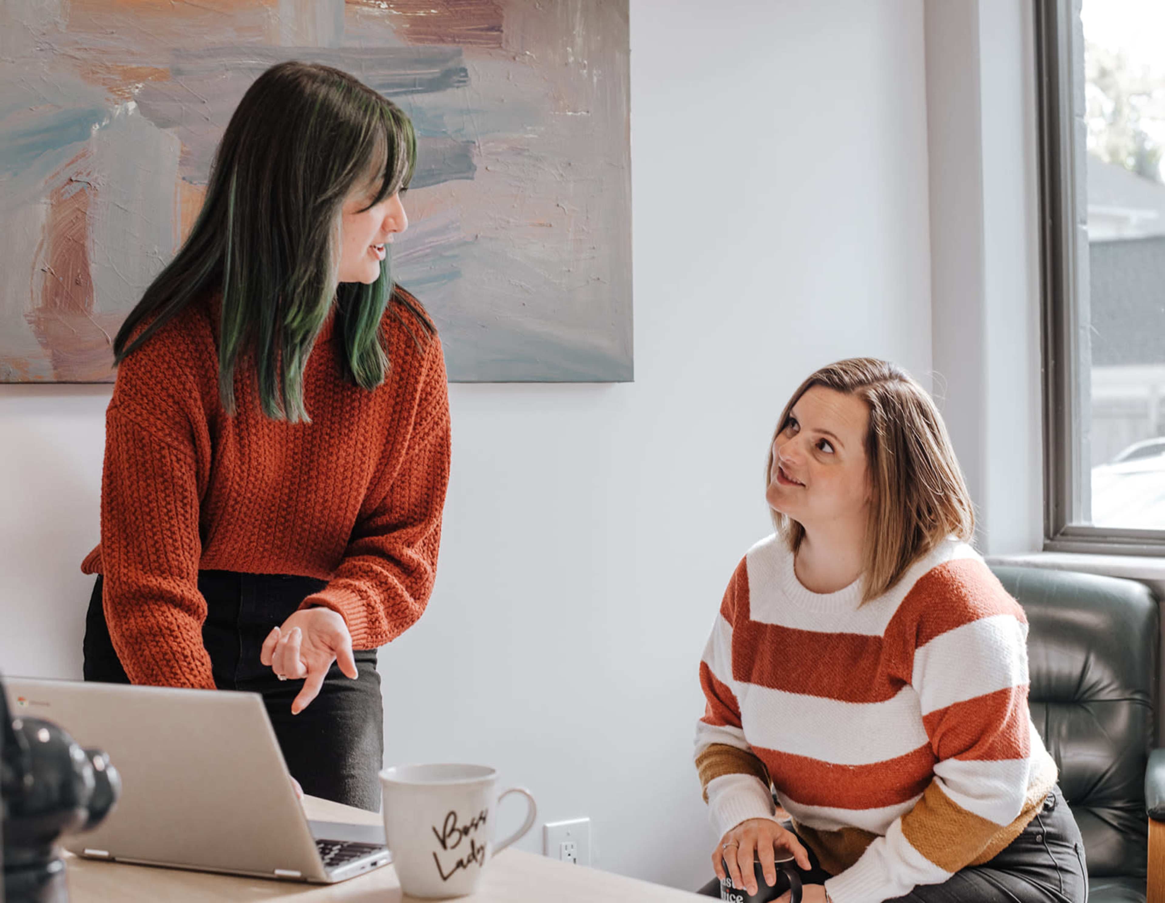 Two women are engaged in a conversation, with one standing and gesturing while the other sits and looks up attentively, next to a laptop in a well-lit, modern room.