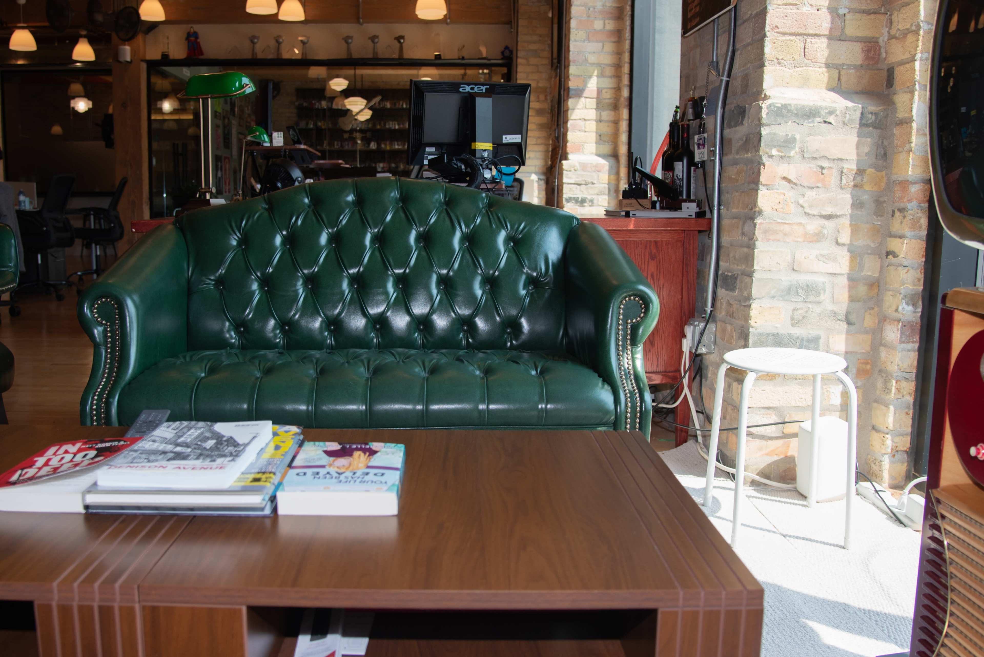 The image features a green leather tufted sofa facing a wooden coffee table, with a computer setup and various items in a well-lit room with brick walls.