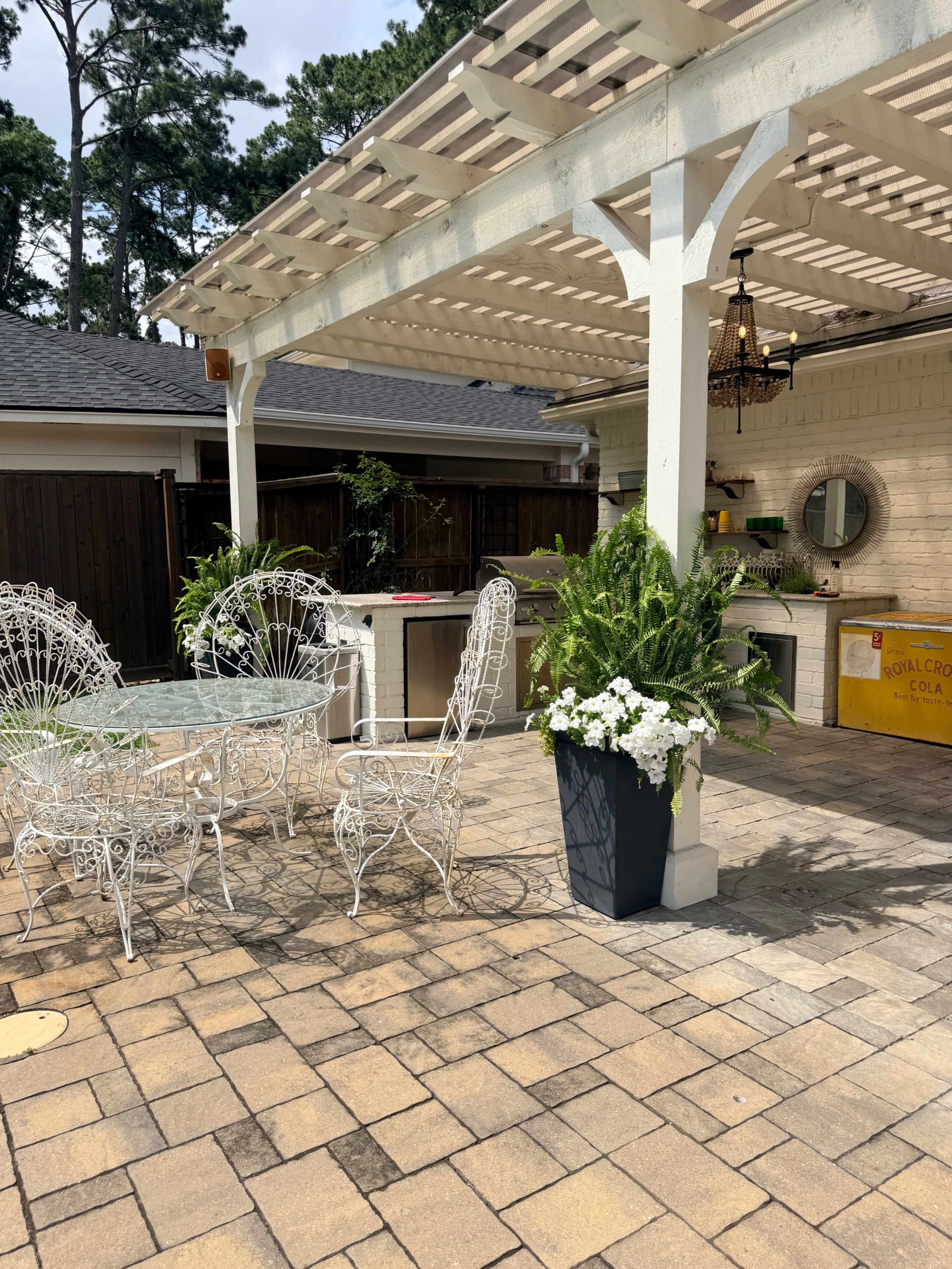 The image shows a shaded outdoor patio with white wrought iron furniture, a stone floor, potted plants, and an outdoor kitchen area under a wooden pergola.