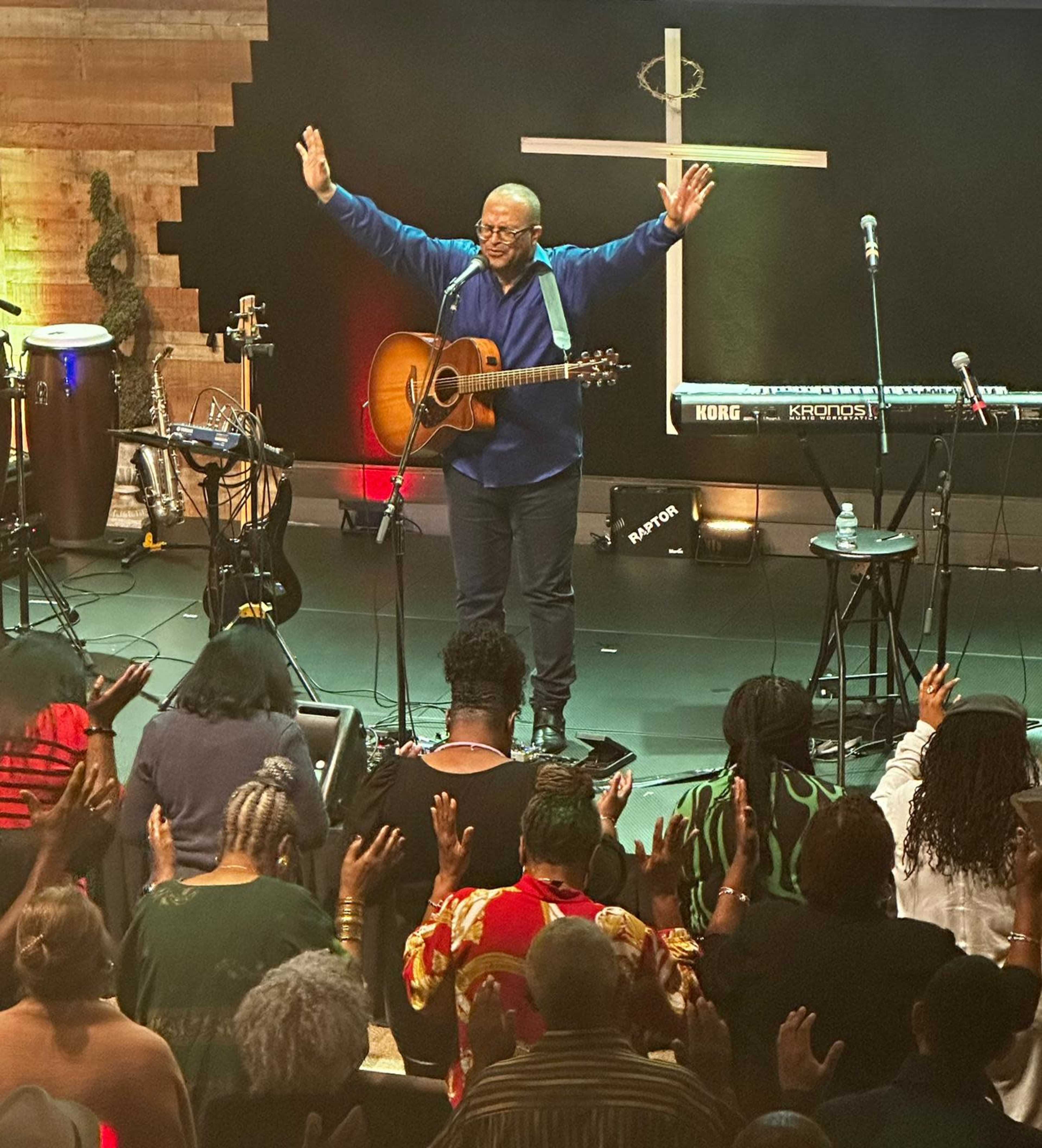 A musician holds a guitar and raises his arms on stage as an audience members worship with hands lifted.