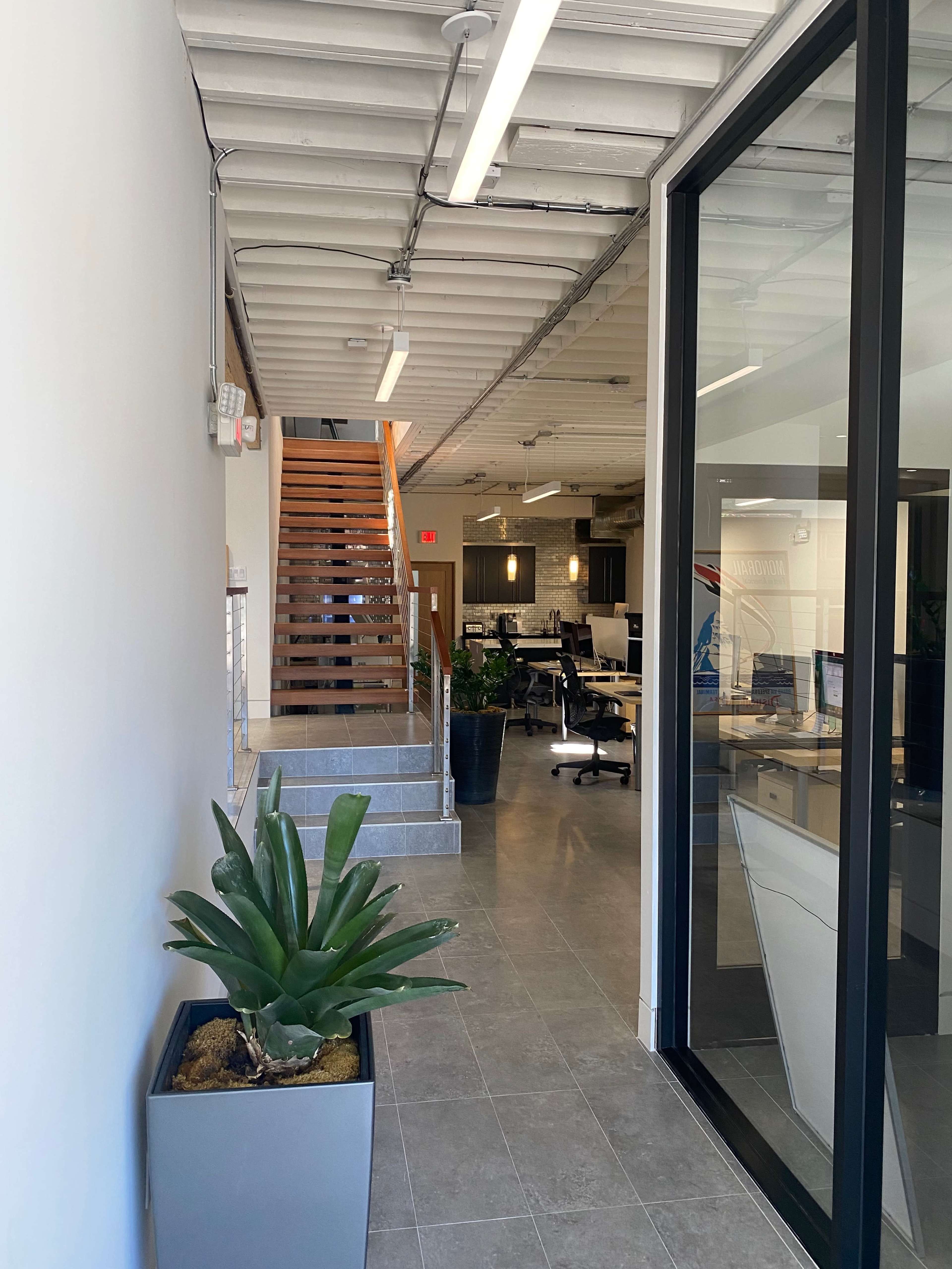 A modern office interior featuring a staircase at the end of a hallway, with plants in planters and workstations visible through large glass panels.