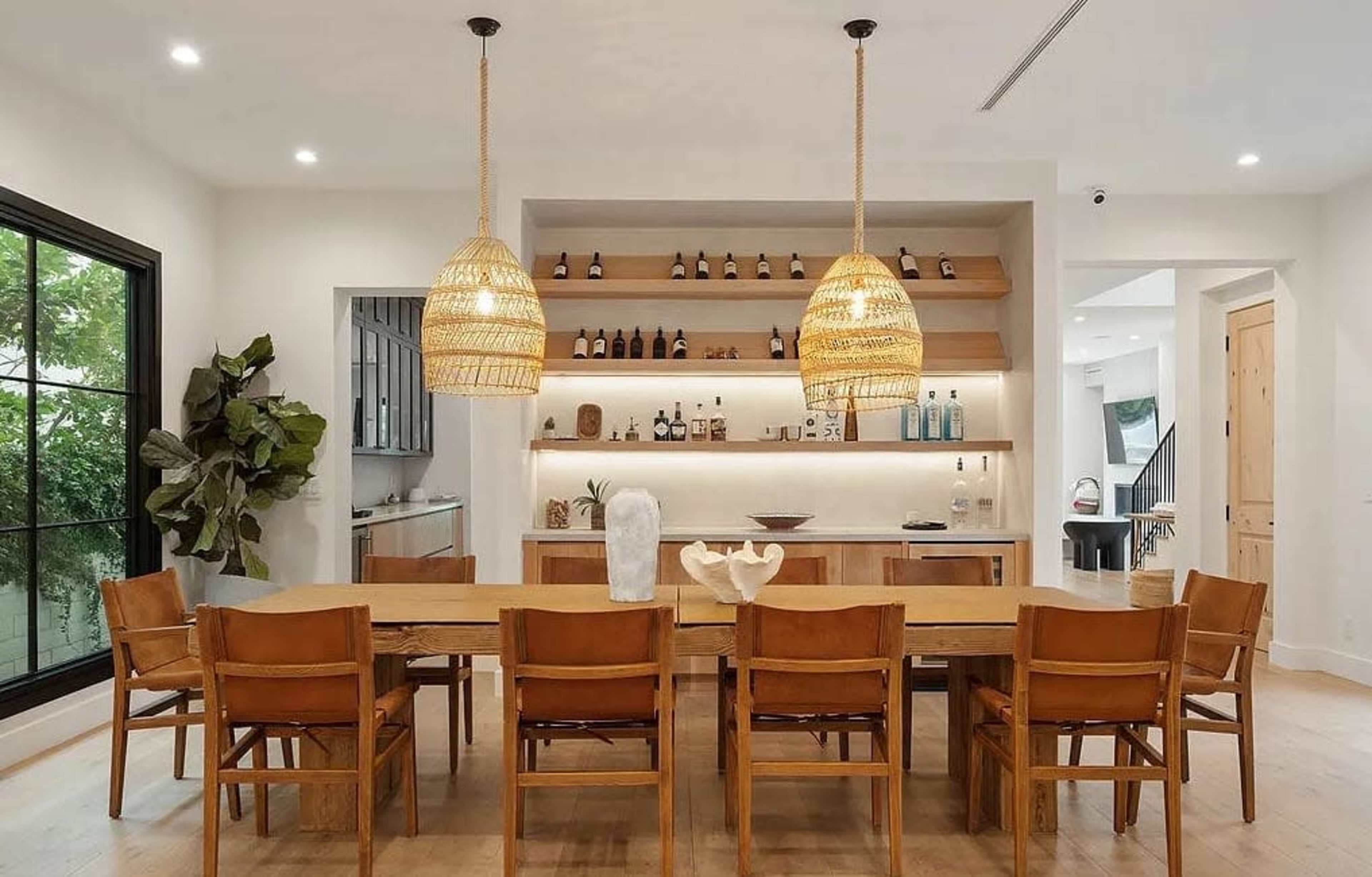 The image shows a modern dining area featuring a long wooden table surrounded by eight leather chairs, with woven pendant lights above and shelves displaying various bottles and decor.