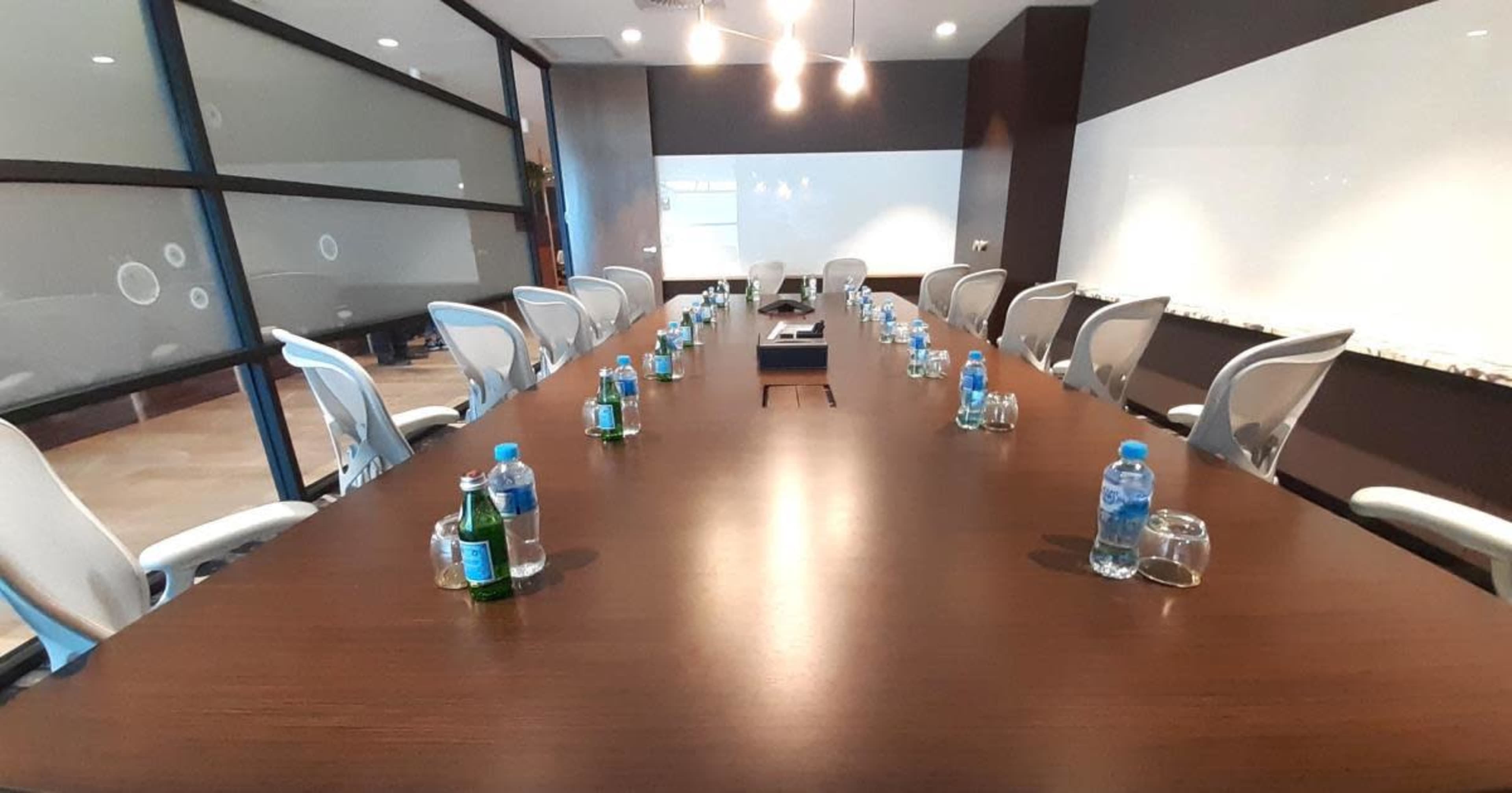 A long, wooden conference table is arranged with water bottles and glasses, surrounded by several chairs in a modern meeting room.