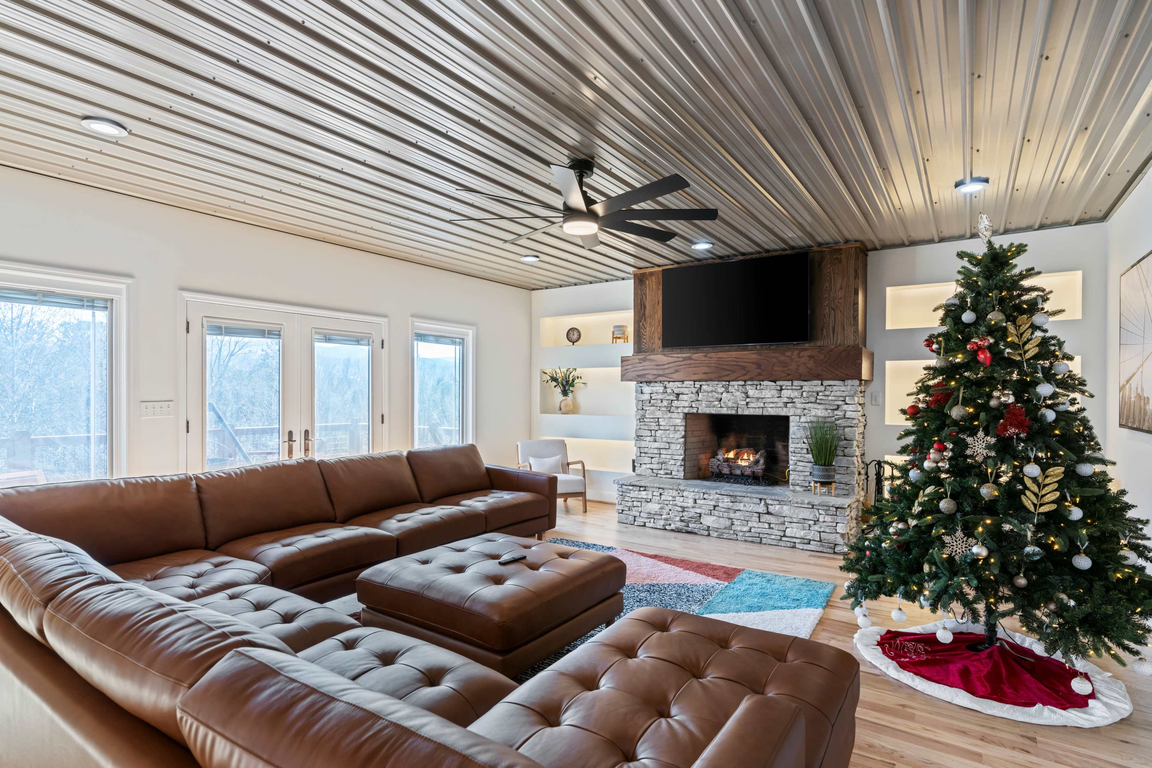 A modern living room features a leather sectional sofa, a stone fireplace with a television above, and a decorated Christmas tree.