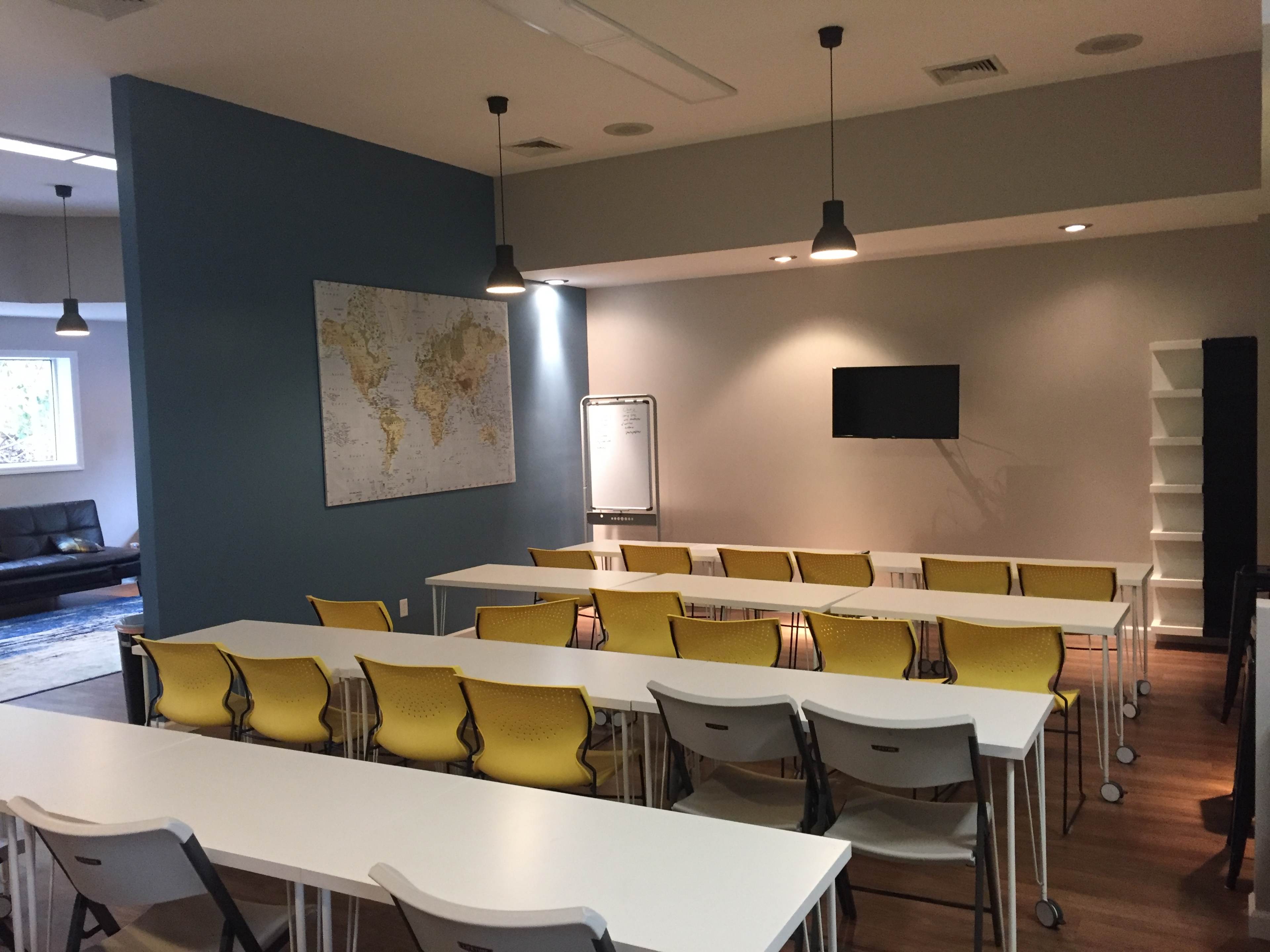 A classroom features yellow chairs and white tables arranged in rows, with a world map on the wall and a television mounted above a whiteboard.