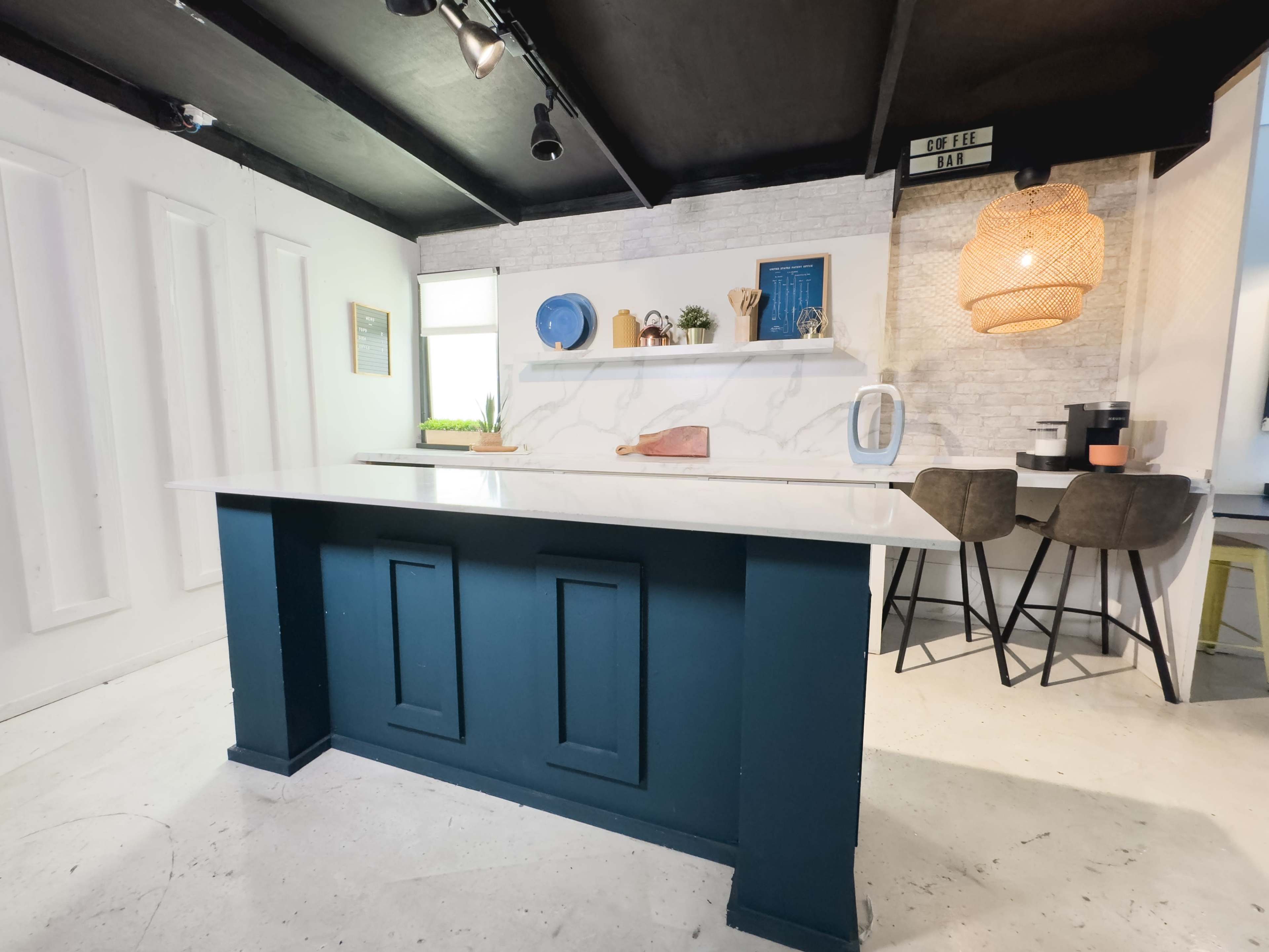 A modern kitchen with a blue island counter, a white brick wall, and a cozy dining area featuring two chairs and decorative items on shelves.