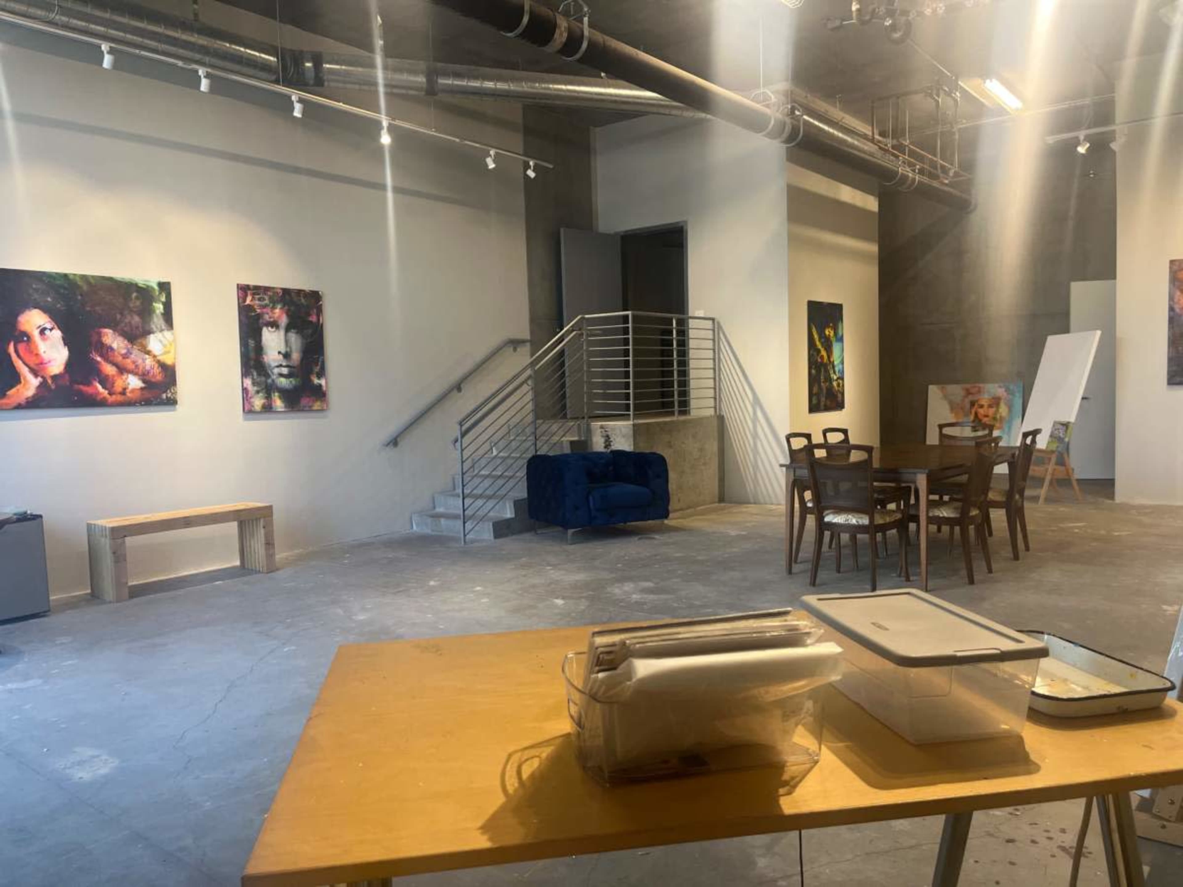 The image shows an art gallery space with paintings on the walls, a dining table and chairs, a couch, and a staircase leading to another level.