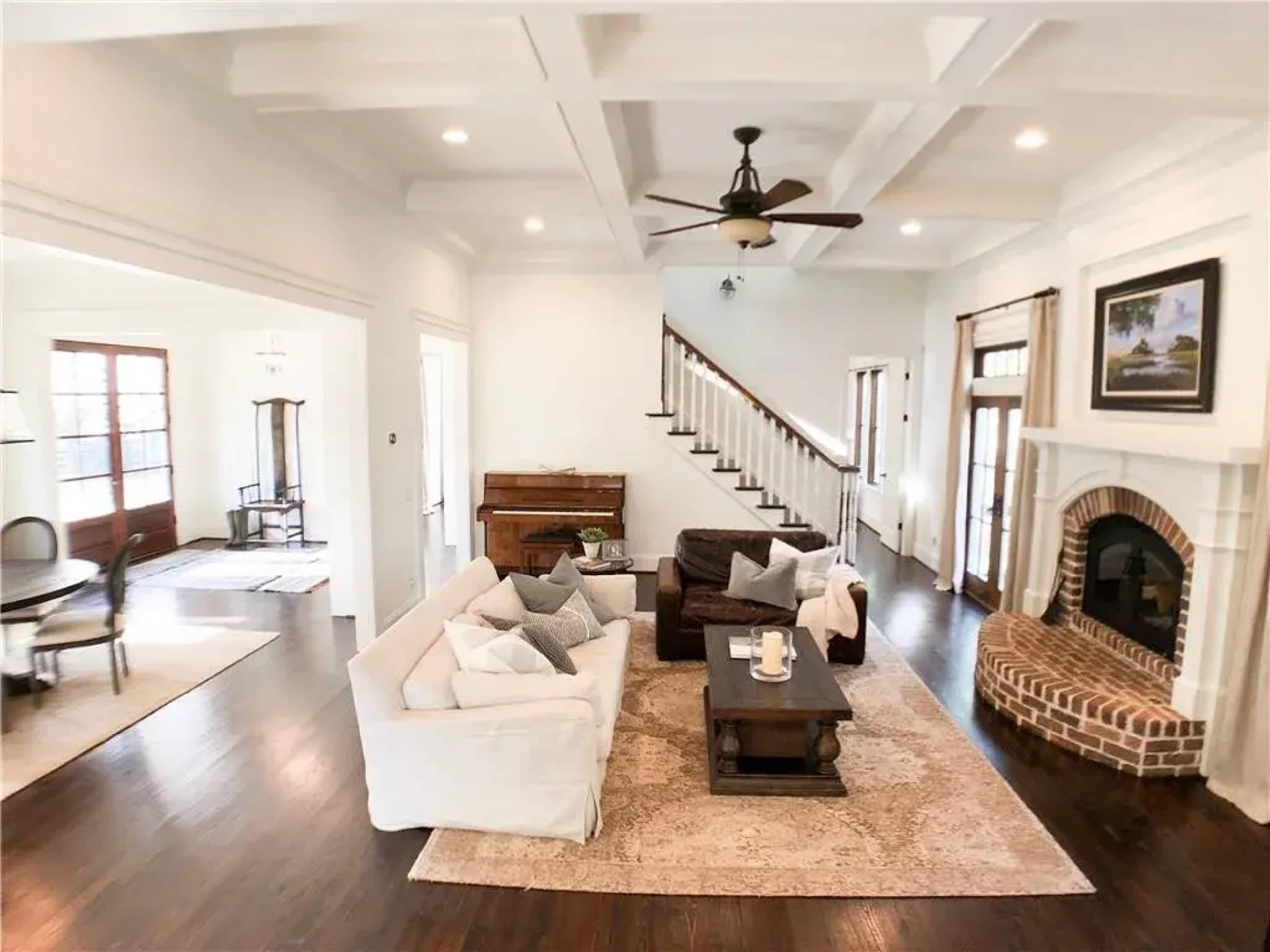 A spacious living room features a white sofa and a brown leather couch surrounding a wooden coffee table, with a staircase and dining area visible in the background.