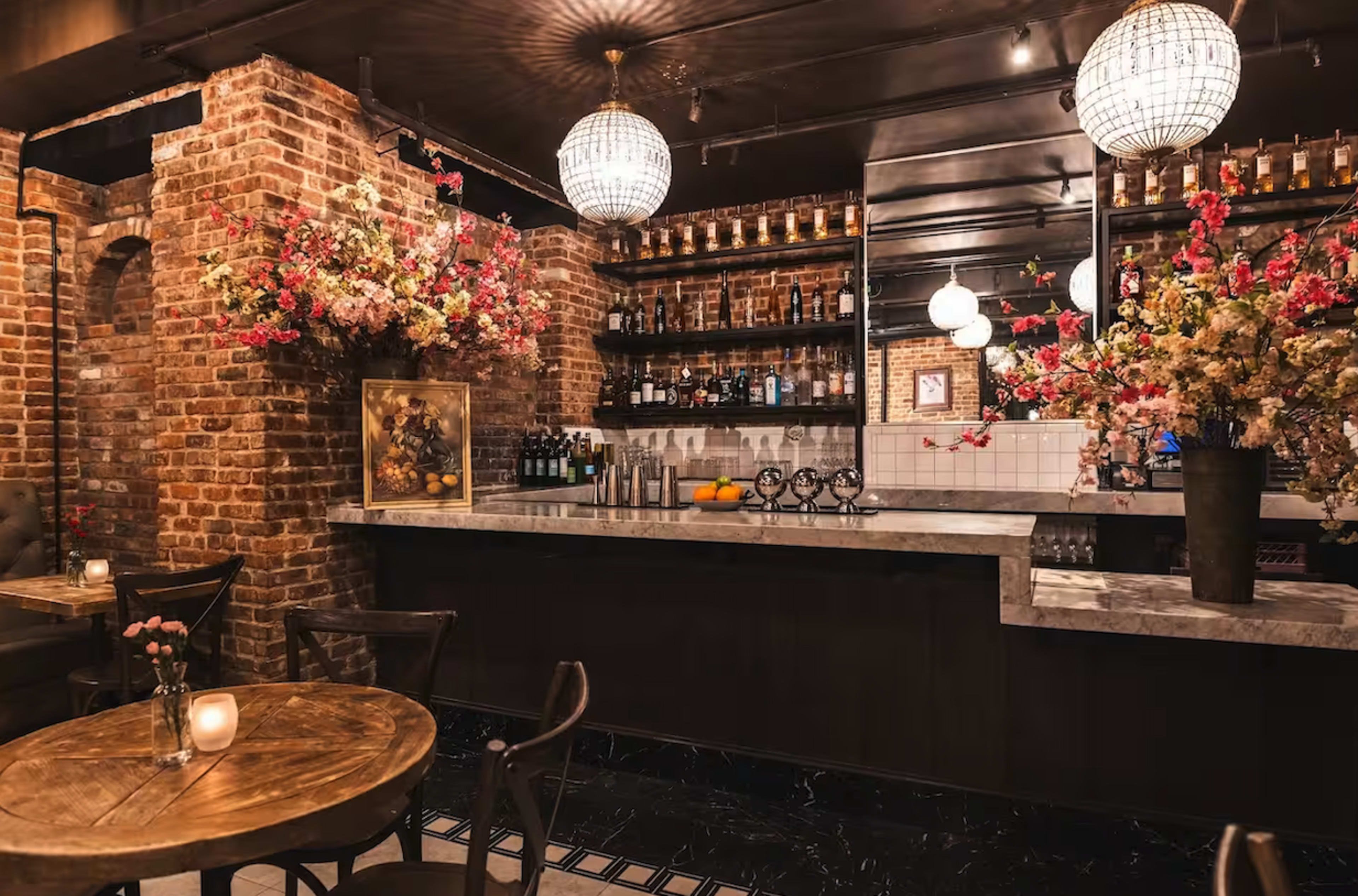 The image shows a stylish bar area with brick walls, marble counters, and floral decorations, illuminated by hanging light fixtures.