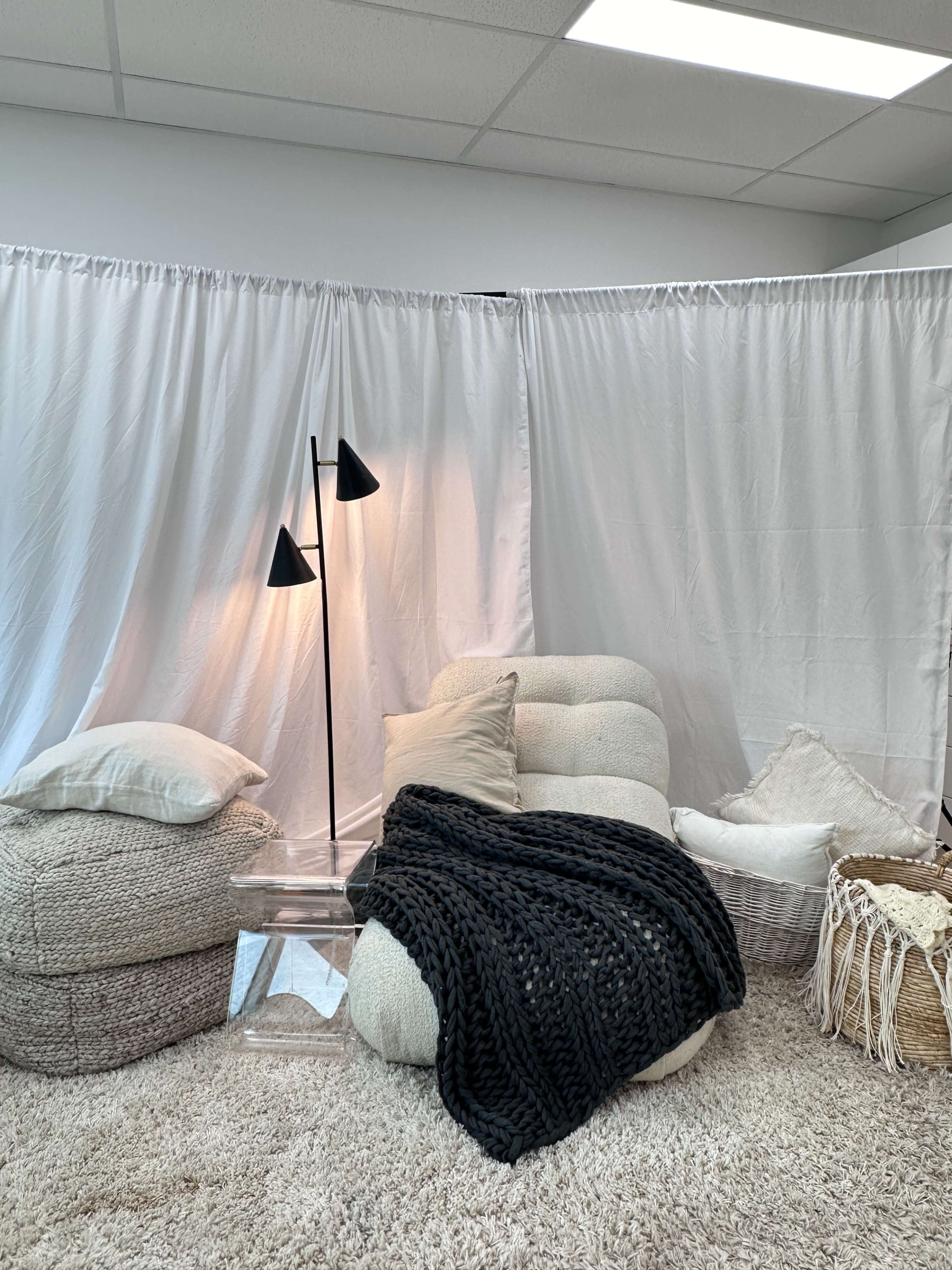 A cozy seating area features textured bean bags and cushions, draped with a knitted throw, next to a black floor lamp on a light carpet.