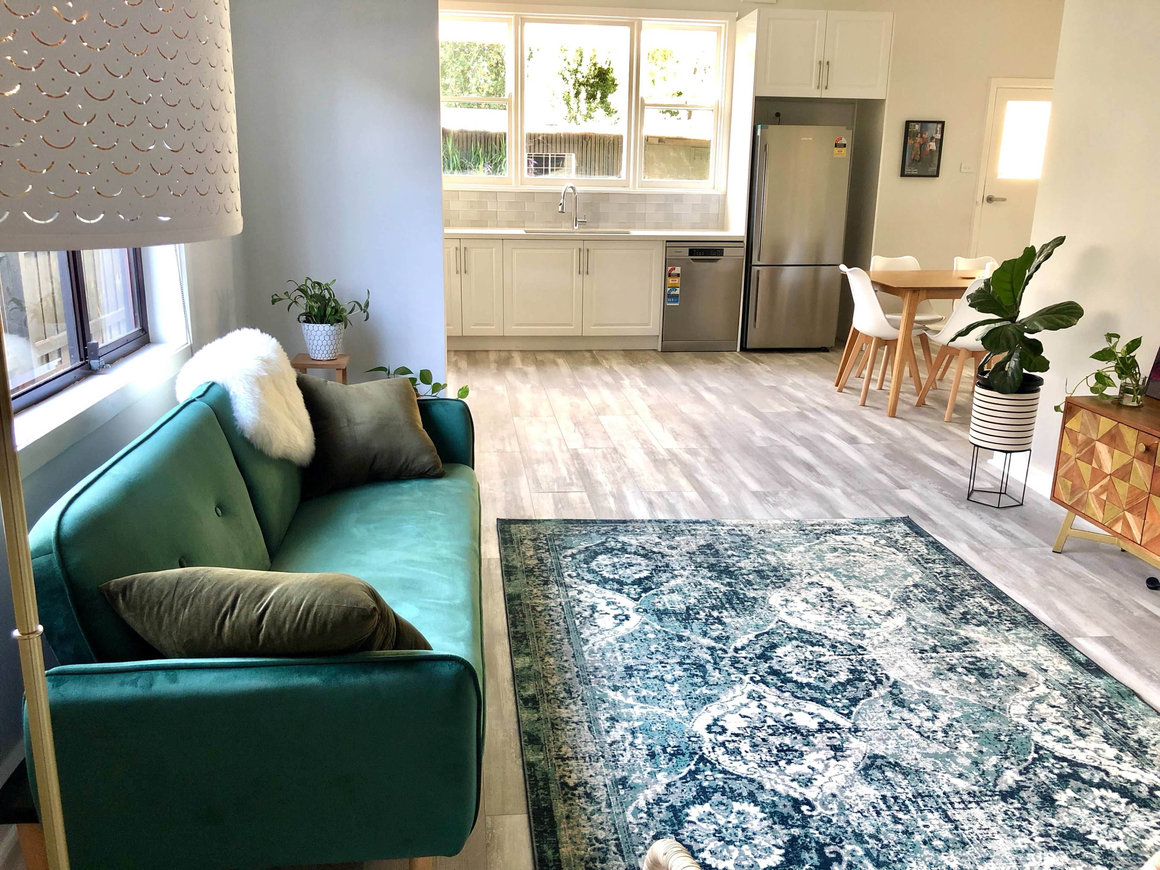 The image shows a modern living space with a green sofa, a patterned blue rug, a dining area with a wooden table and chairs, and a kitchen featuring white cabinetry and stainless steel appliances.