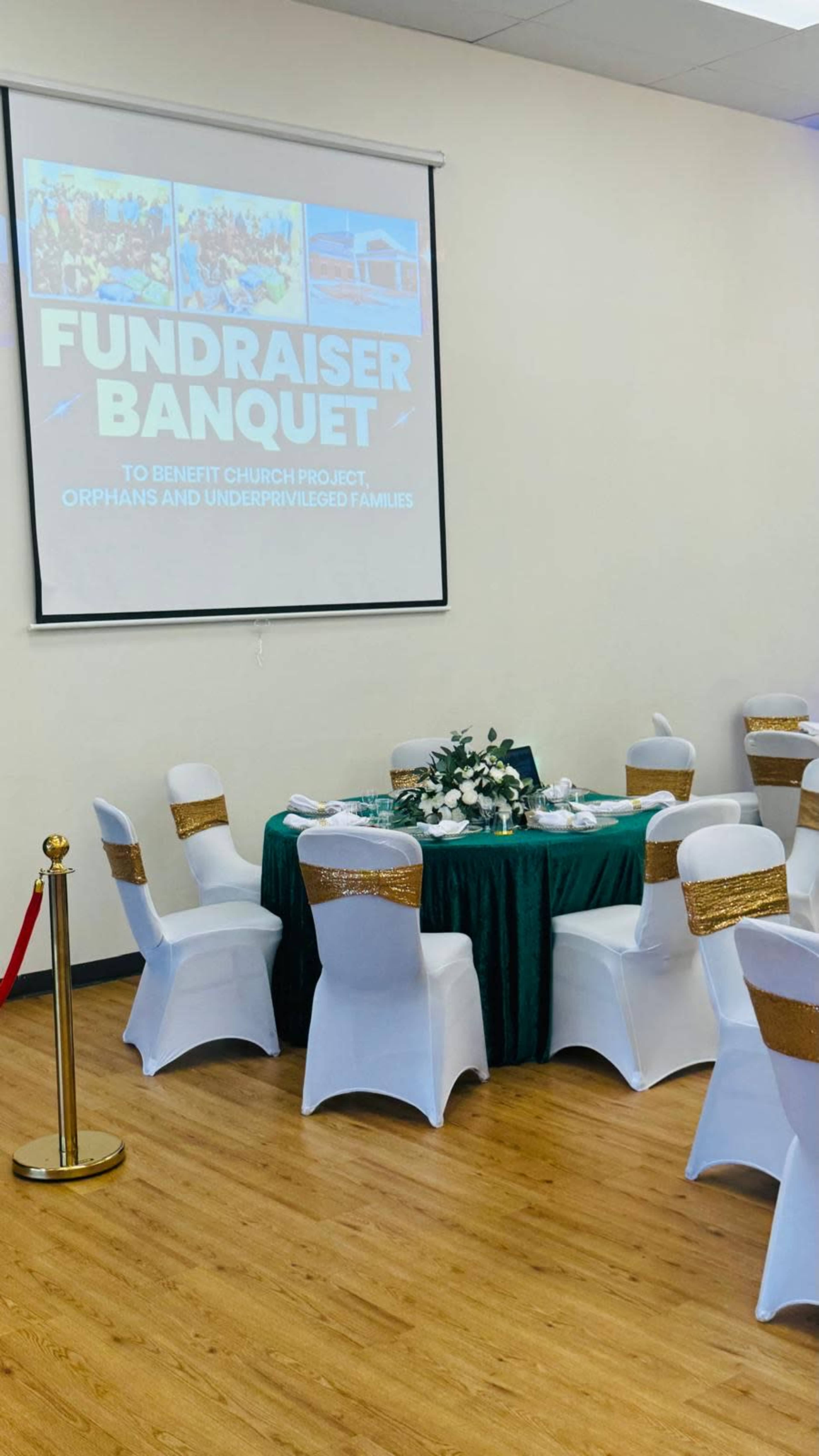 The image shows a banquet setup with a green tablecloth and white chairs adorned with gold sashes, facing a screen displaying information about a fundraiser for orphans and underprivileged families.