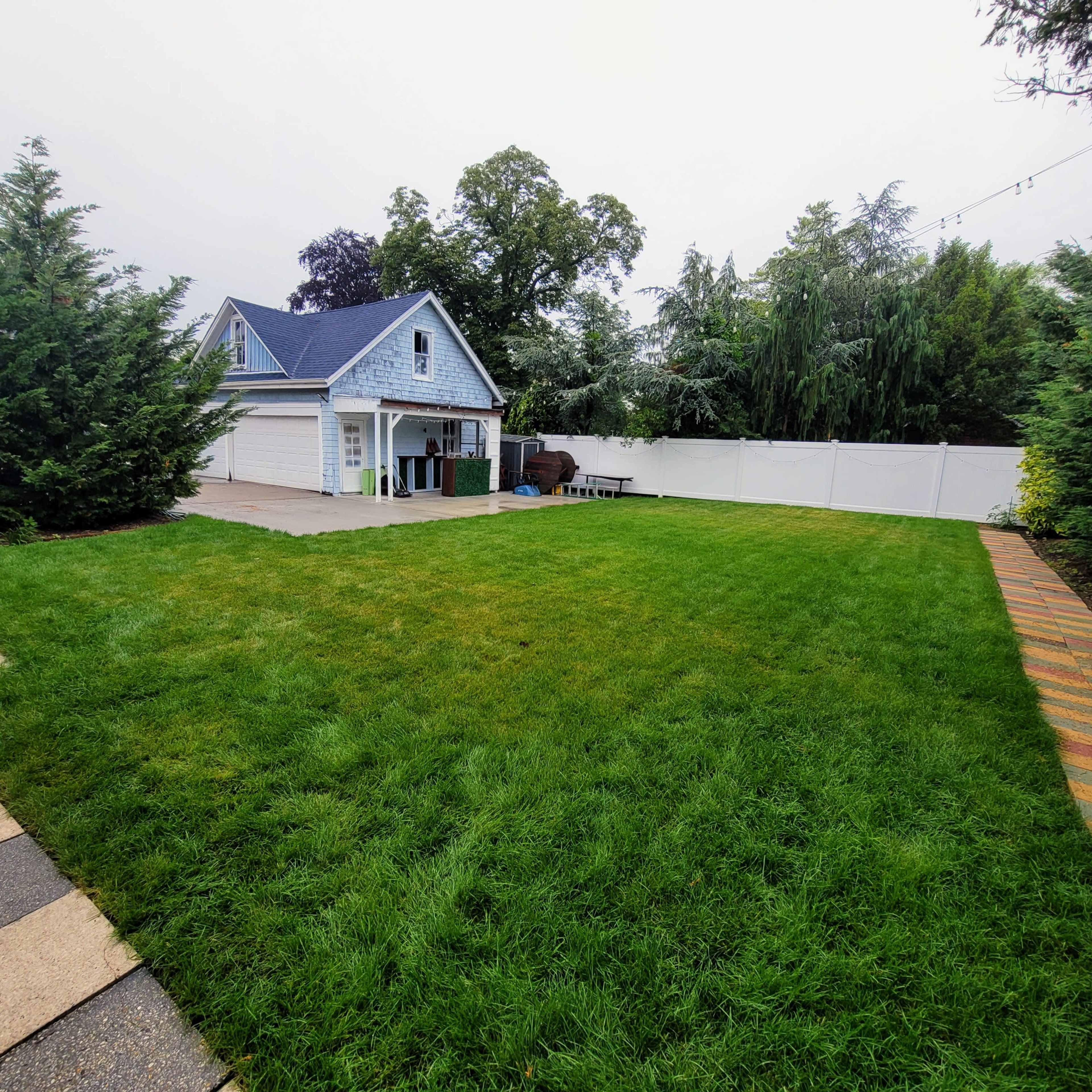 The image shows a spacious backyard with a well-maintained lawn, a wooden patio area, and a small blue shed in the background.