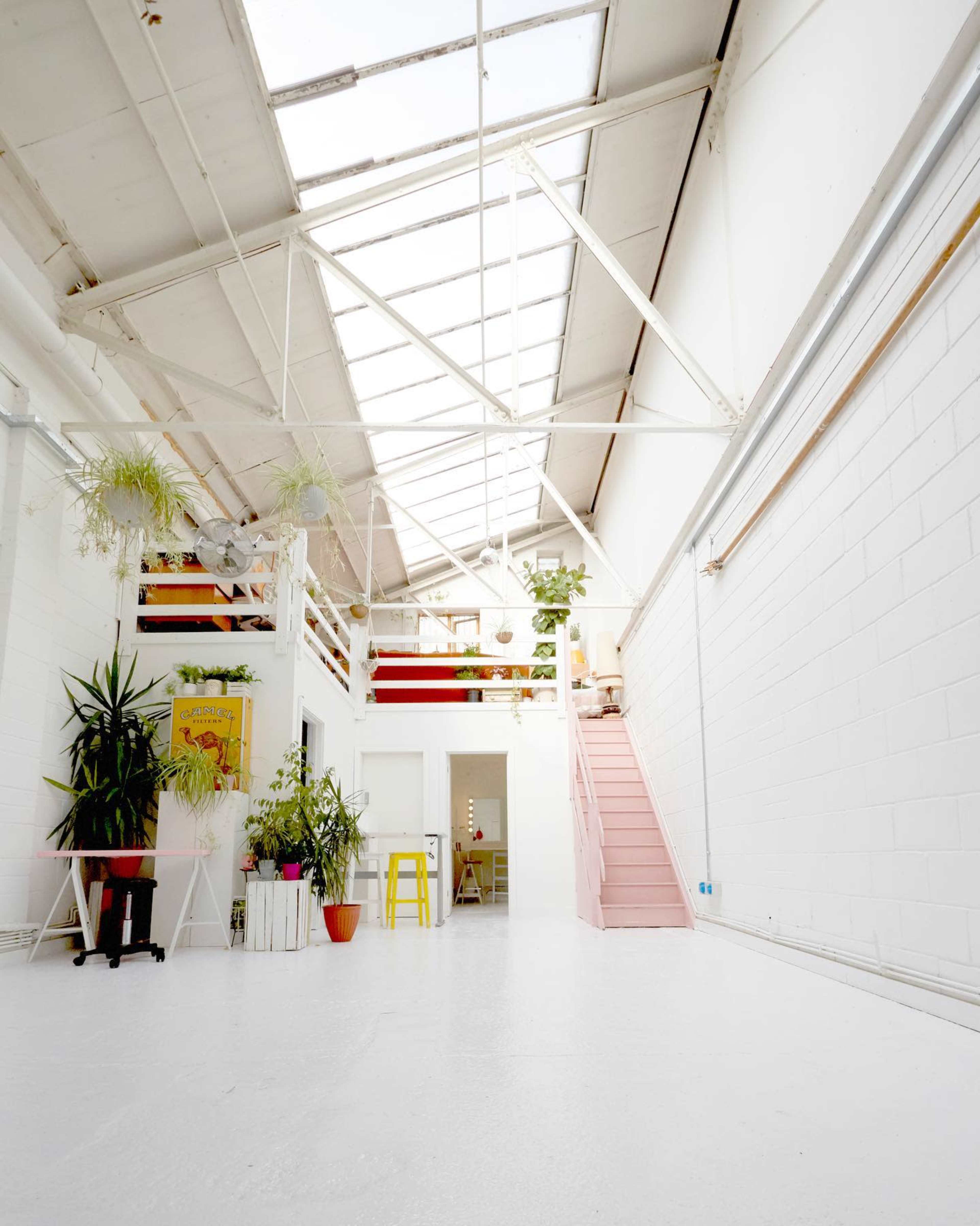 A spacious, industrial-style interior with a high ceiling, large skylights, and a staircase leading to a loft area surrounded by various plants.