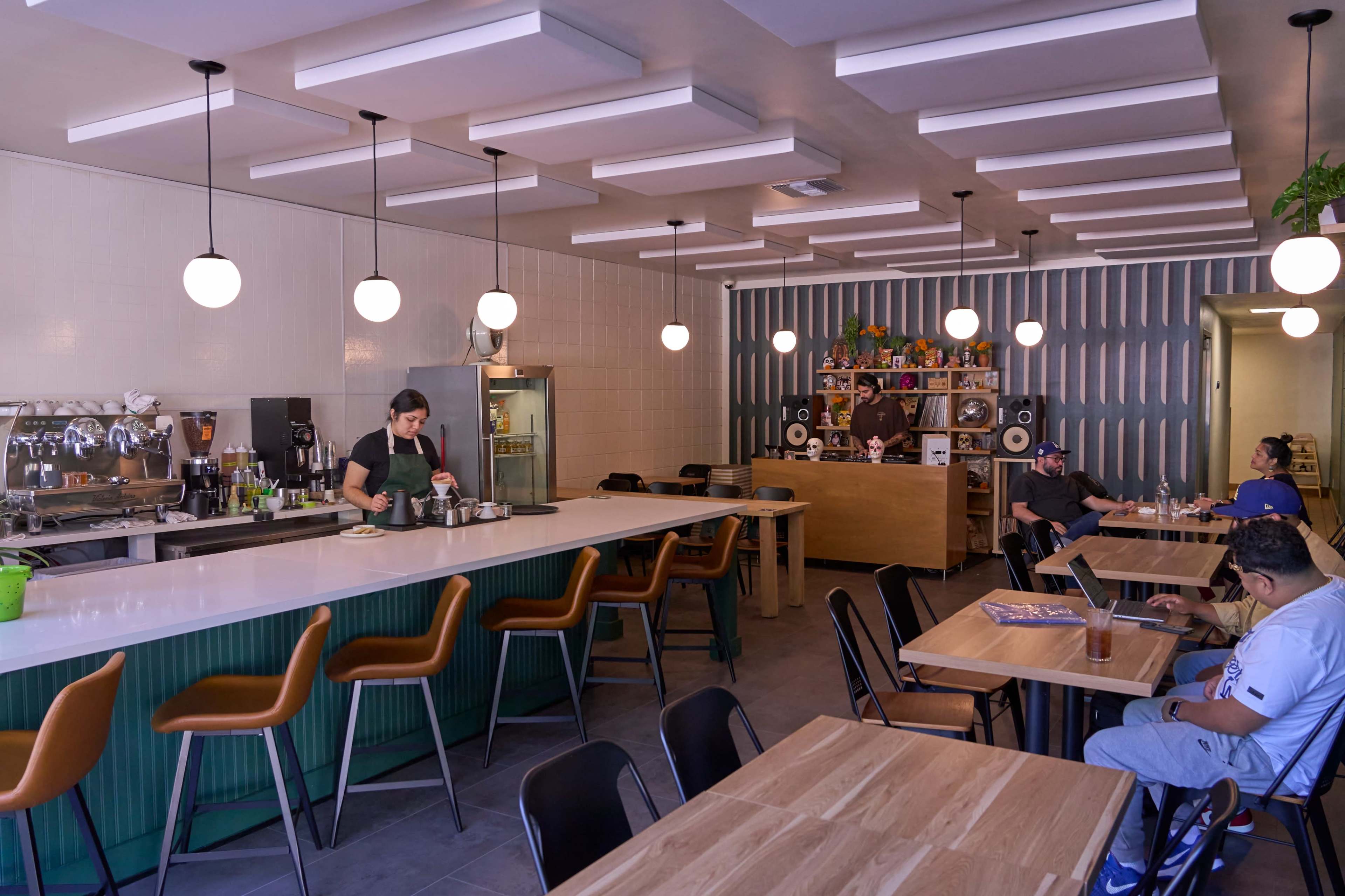 A modern café features a sleek counter with a barista preparing drinks, while patrons sit at wooden tables and a well-stocked bar is visible in the background.