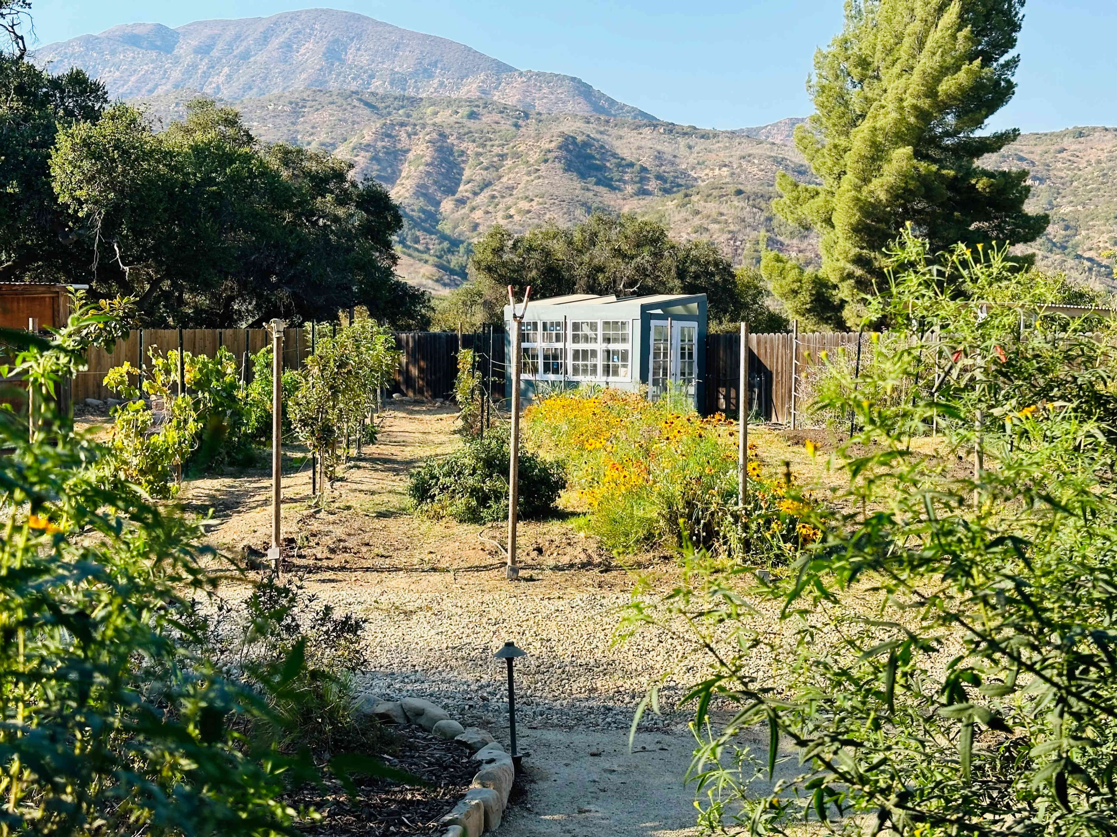 A garden path leads through a cultivated area with flowering plants and a greenhouse, set against a backdrop of rolling mountains.