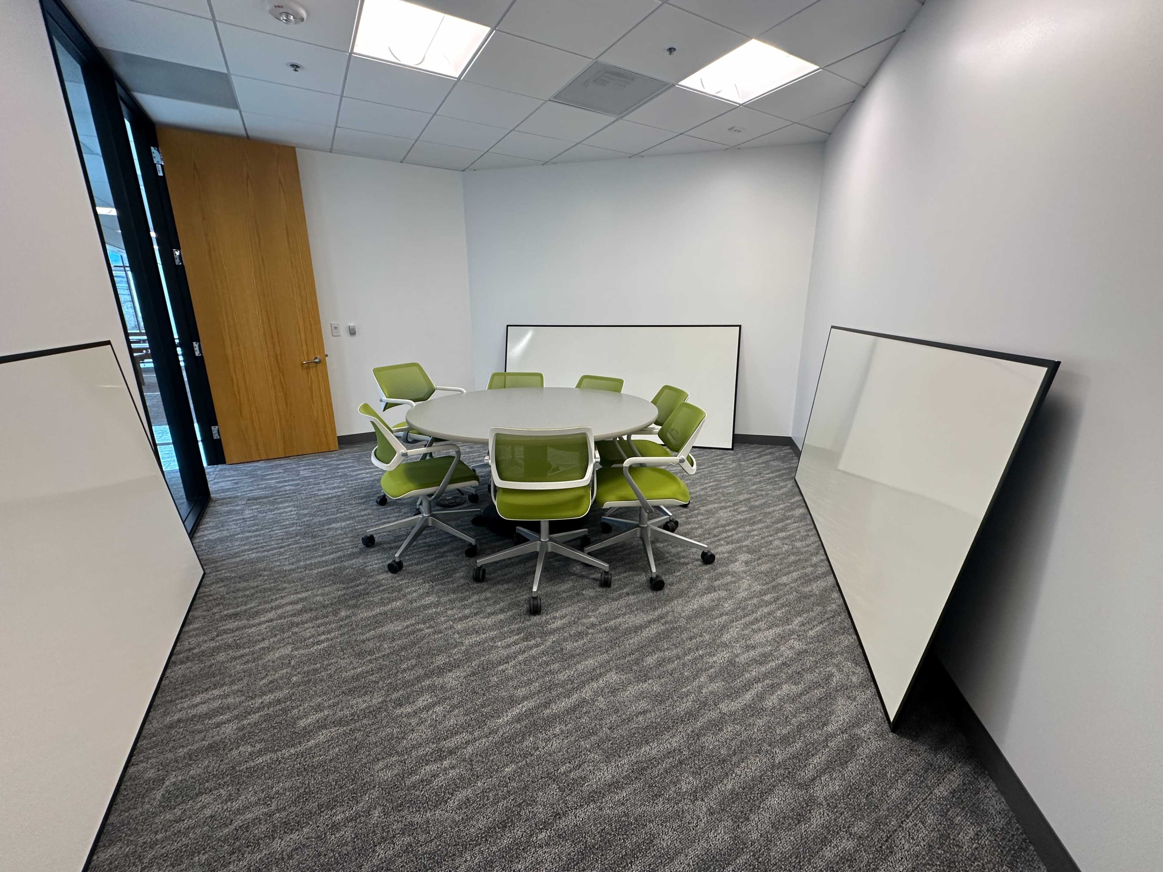 A small conference room features a round table surrounded by green chairs, with two large white boards leaning against the walls.
