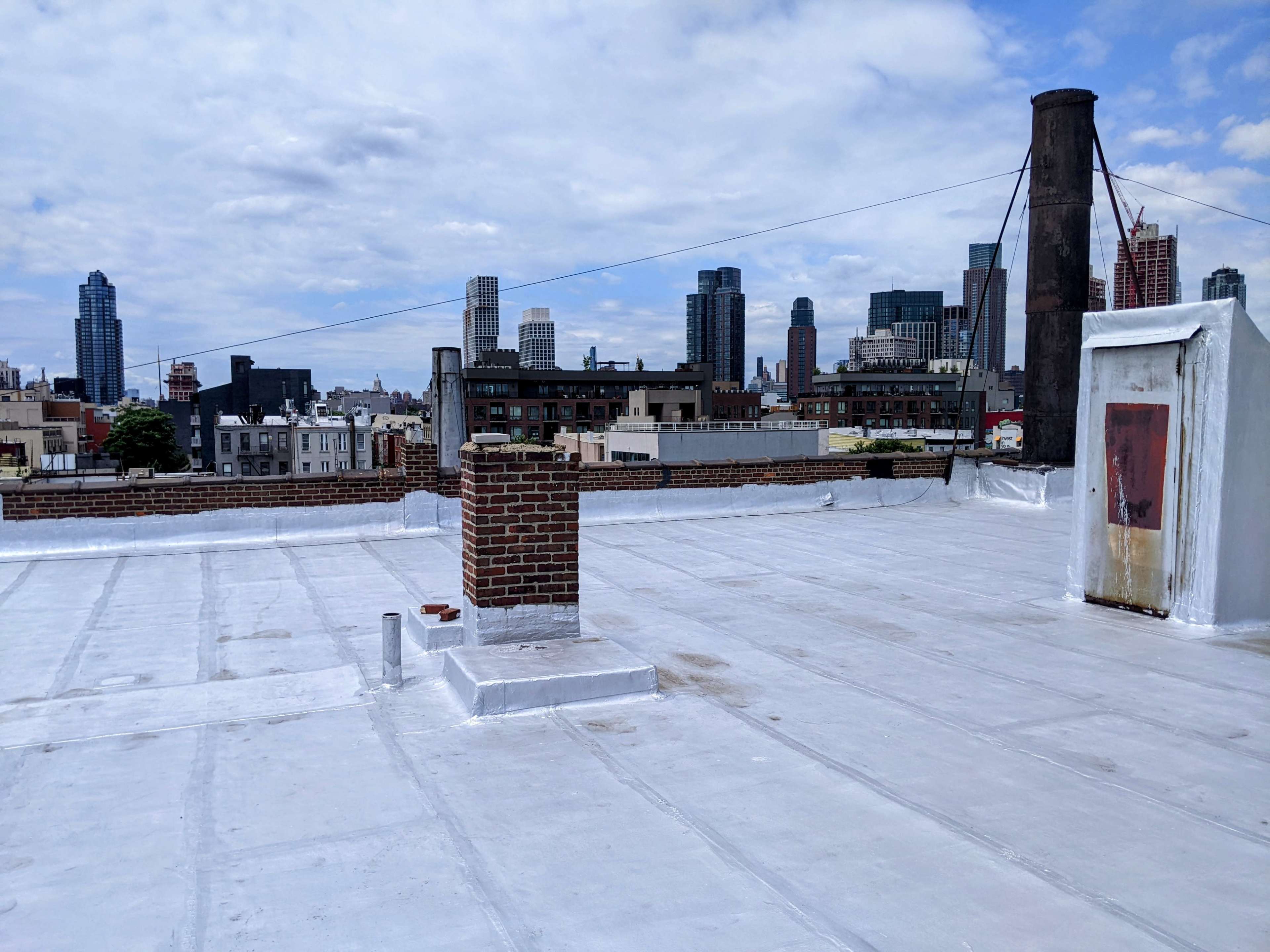 A flat, white rooftop with a brick chimney, overlooking a city skyline with various tall buildings.