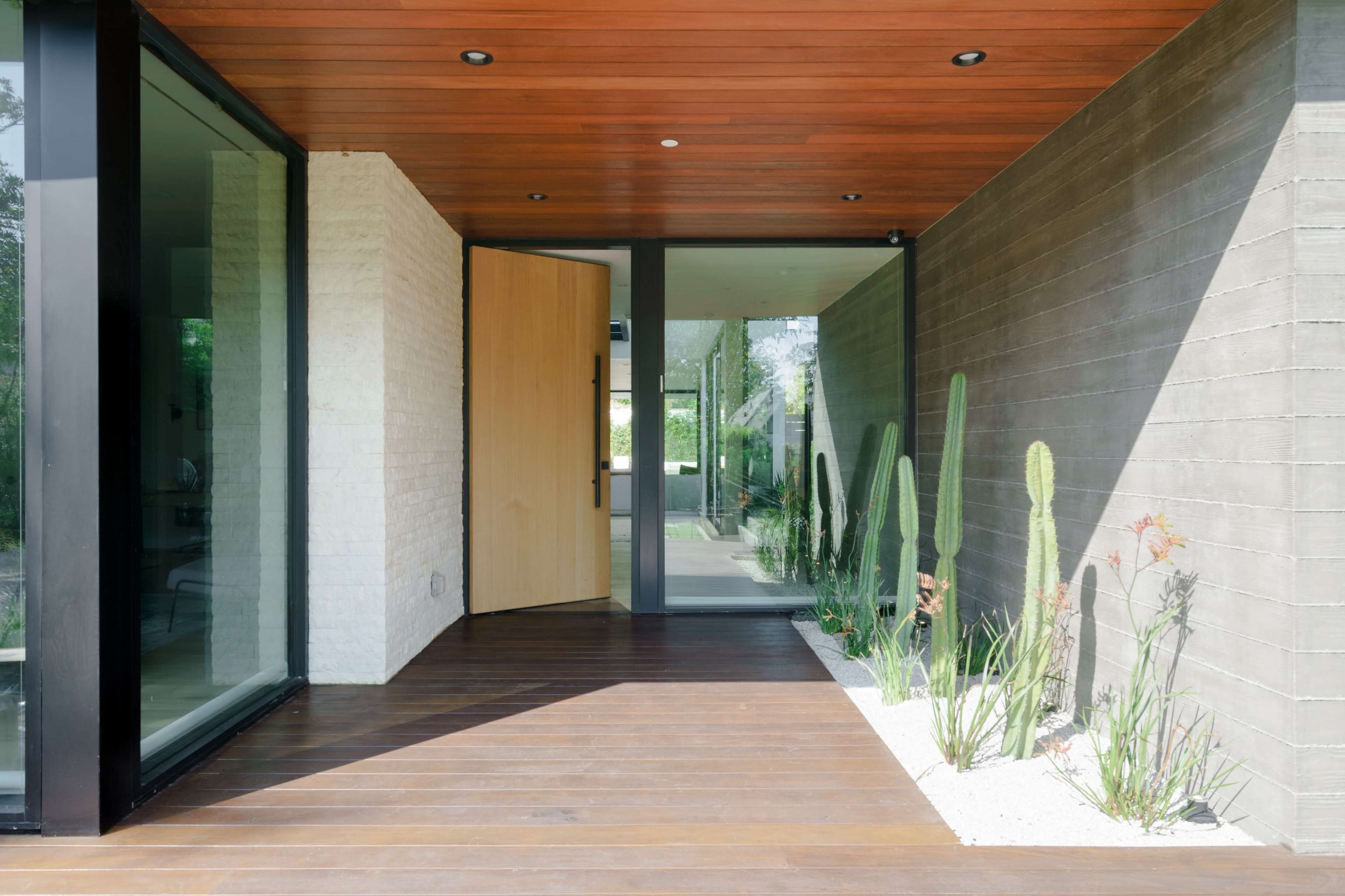 An entrance area featuring a wooden door, large glass windows, and a landscaped space with vertical cacti and other plants on a wooden floor.