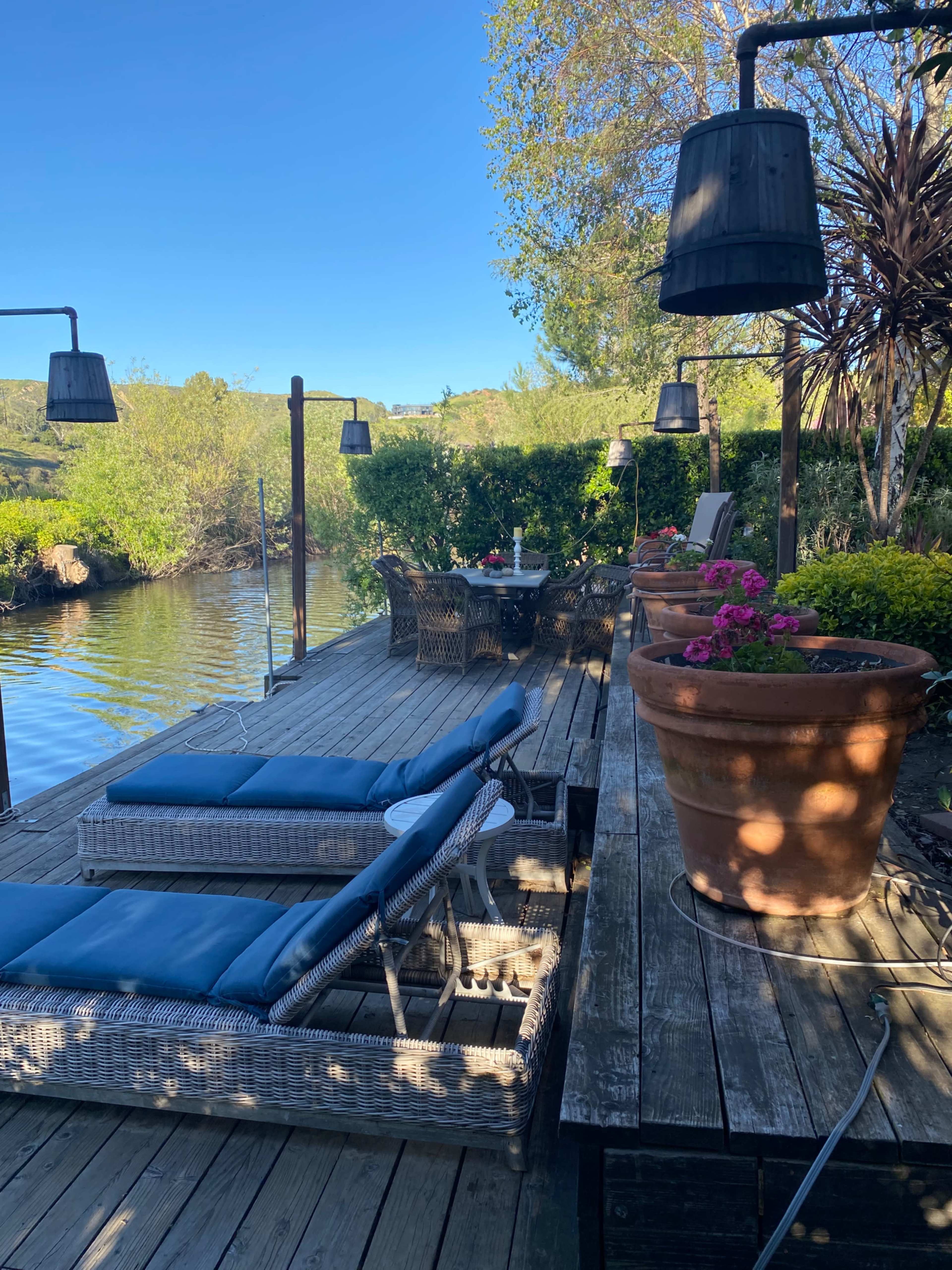 The image shows a wooden dock by a river, featuring blue lounge chairs, potted plants, and outdoor lighting fixtures.
