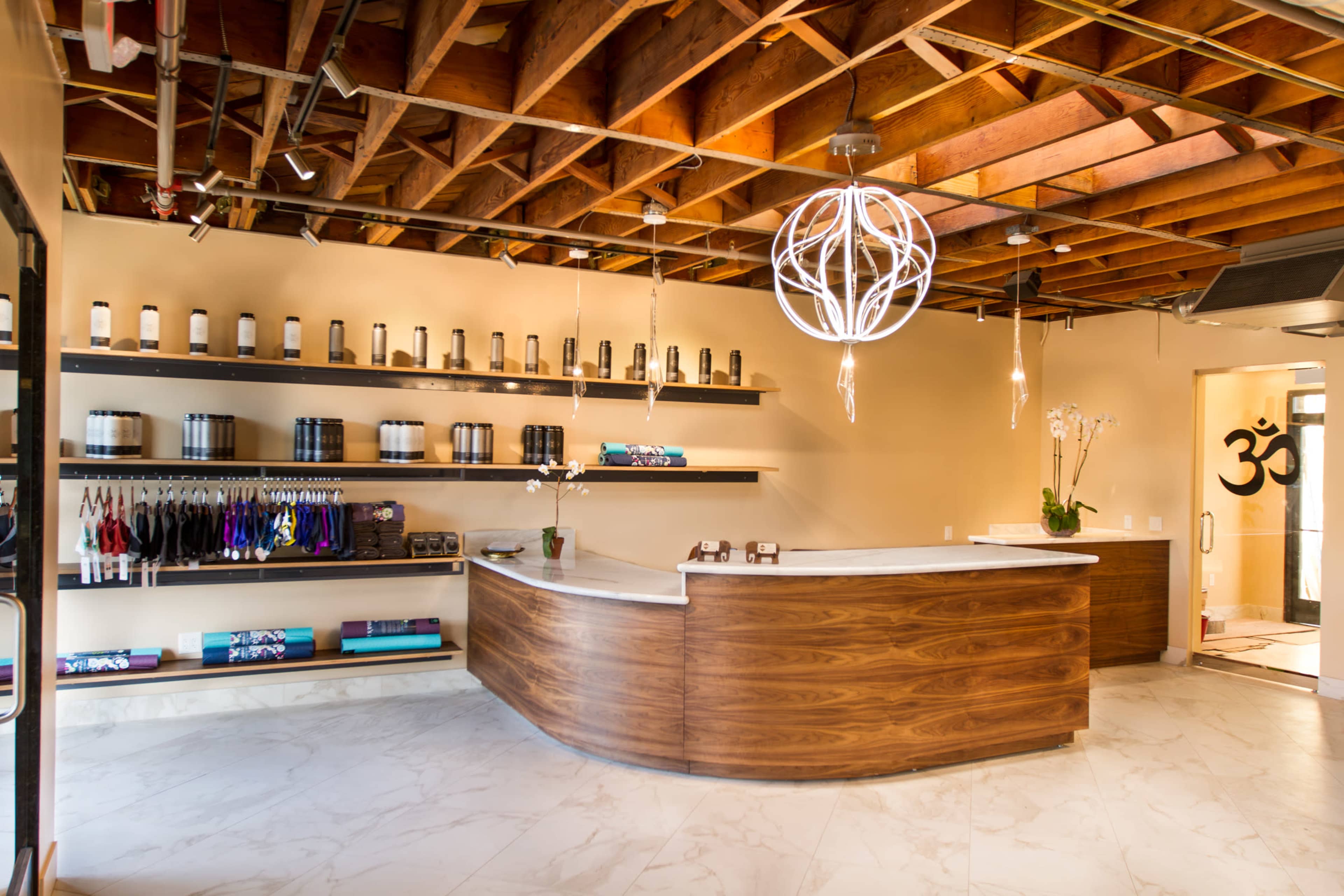The image shows a modern reception area with a curved wooden front desk, shelving displaying various products, and a large decorative light fixture hanging from the ceiling.