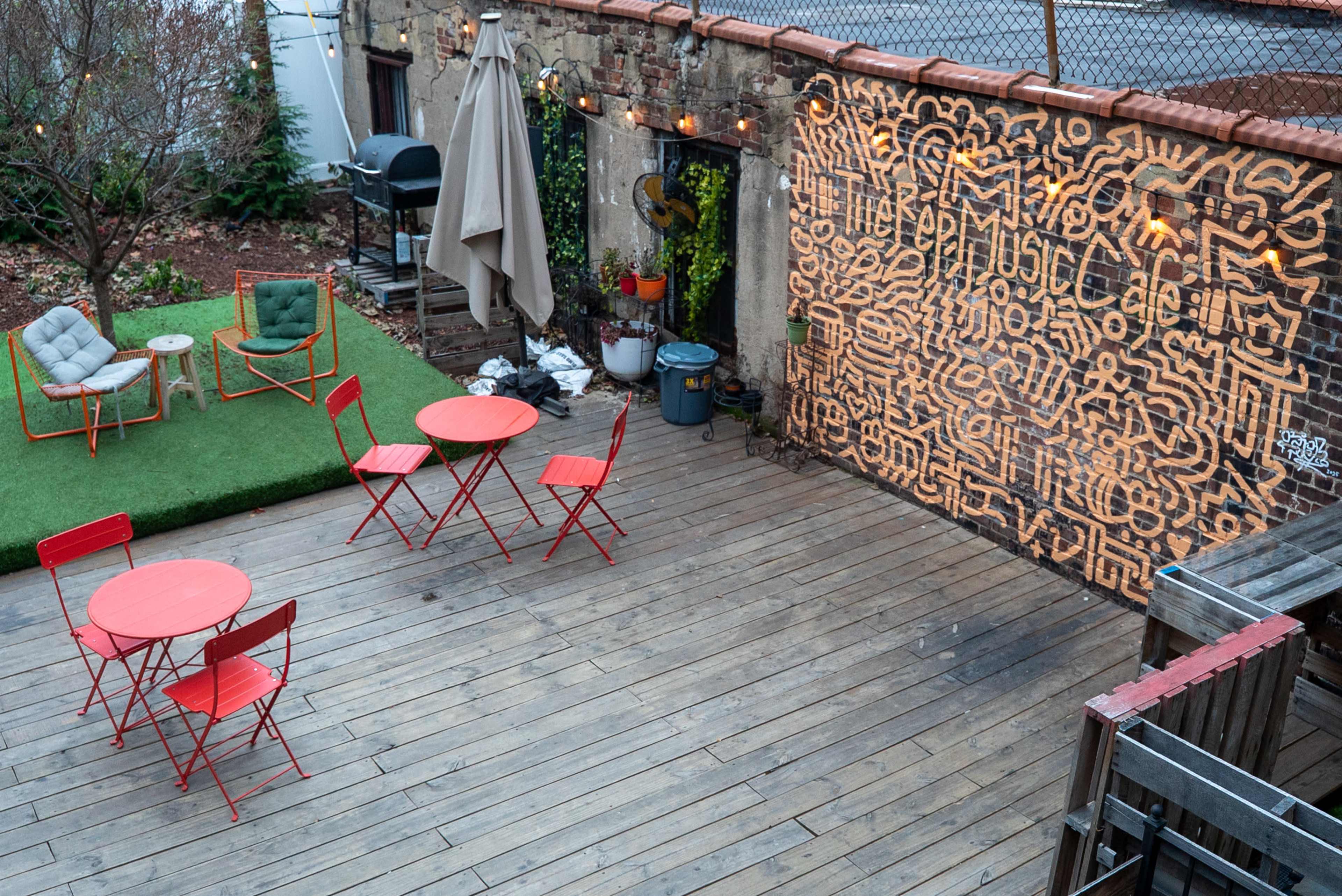The image shows an outdoor patio with wooden flooring and several red tables and chairs, alongside a green grassy area and a patterned wall.