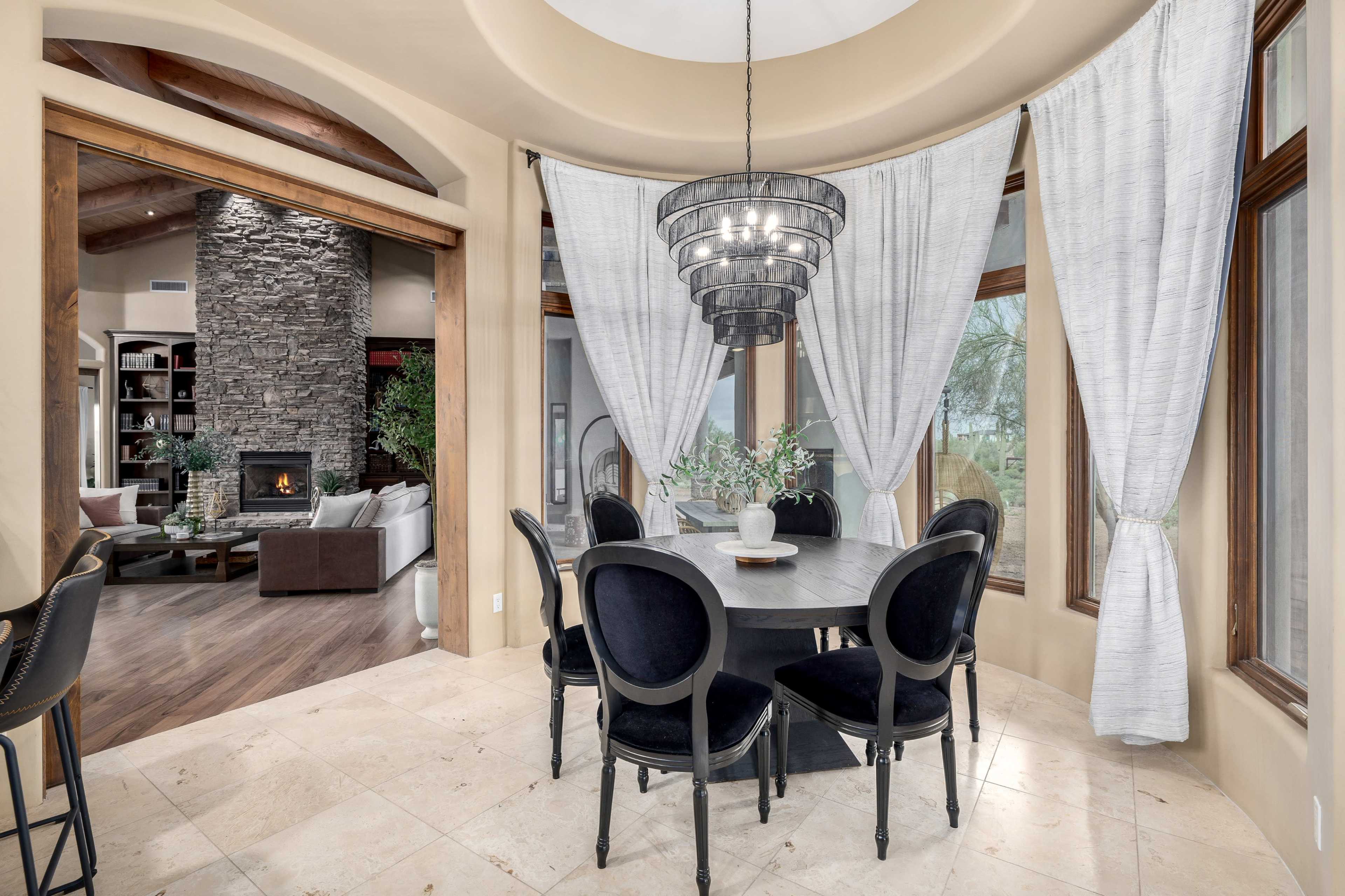 A round dining table with black chairs is surrounded by large windows, offering a view of a living area with a fireplace and a stone accent wall.