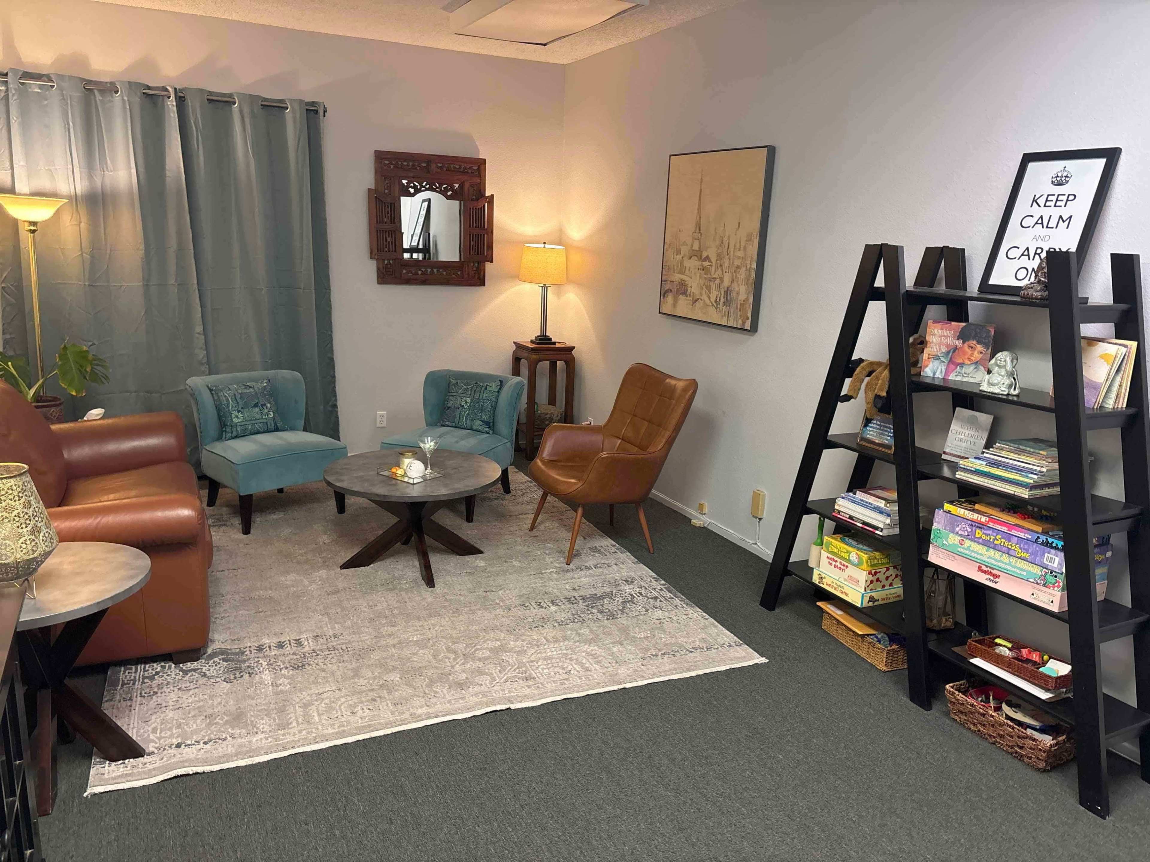 The image depicts a cozy sitting area furnished with two blue armchairs, a brown leather chair, a coffee table, and a bookshelf, all arranged around a light-colored area rug.