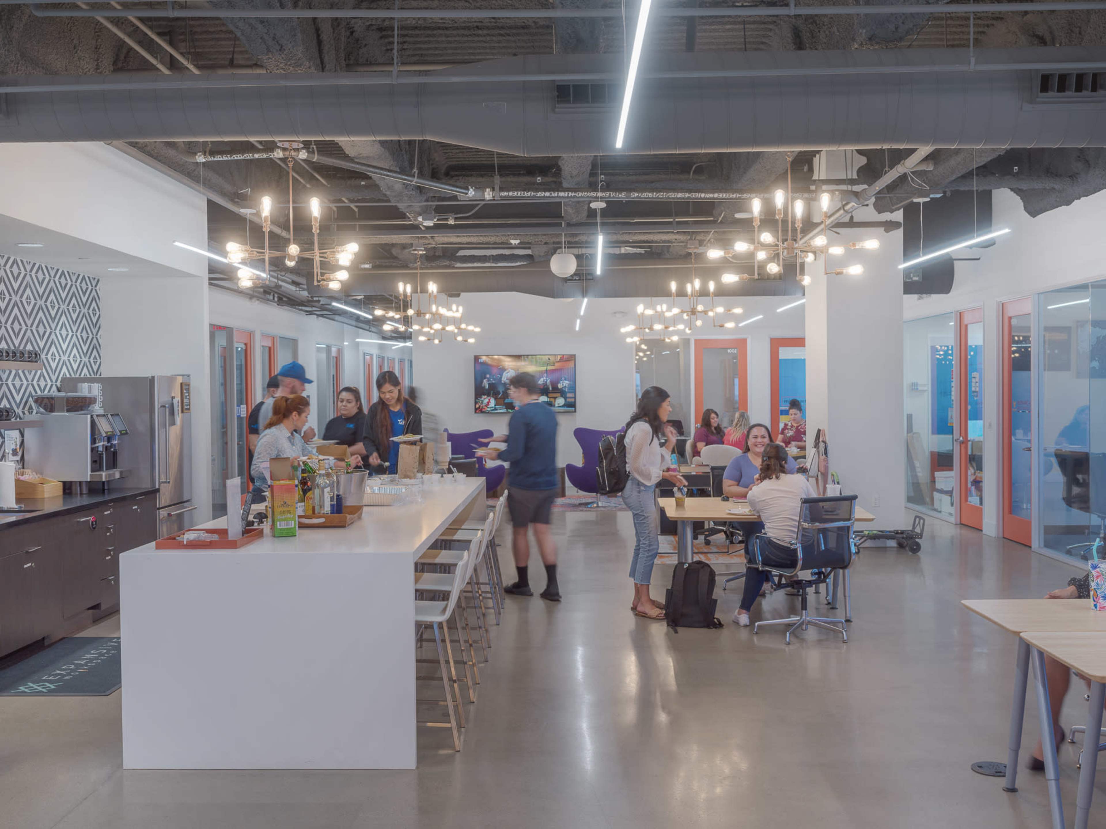 The image shows a modern office space with a kitchen area, collaborative workstations, and a group of people engaging in various activities.