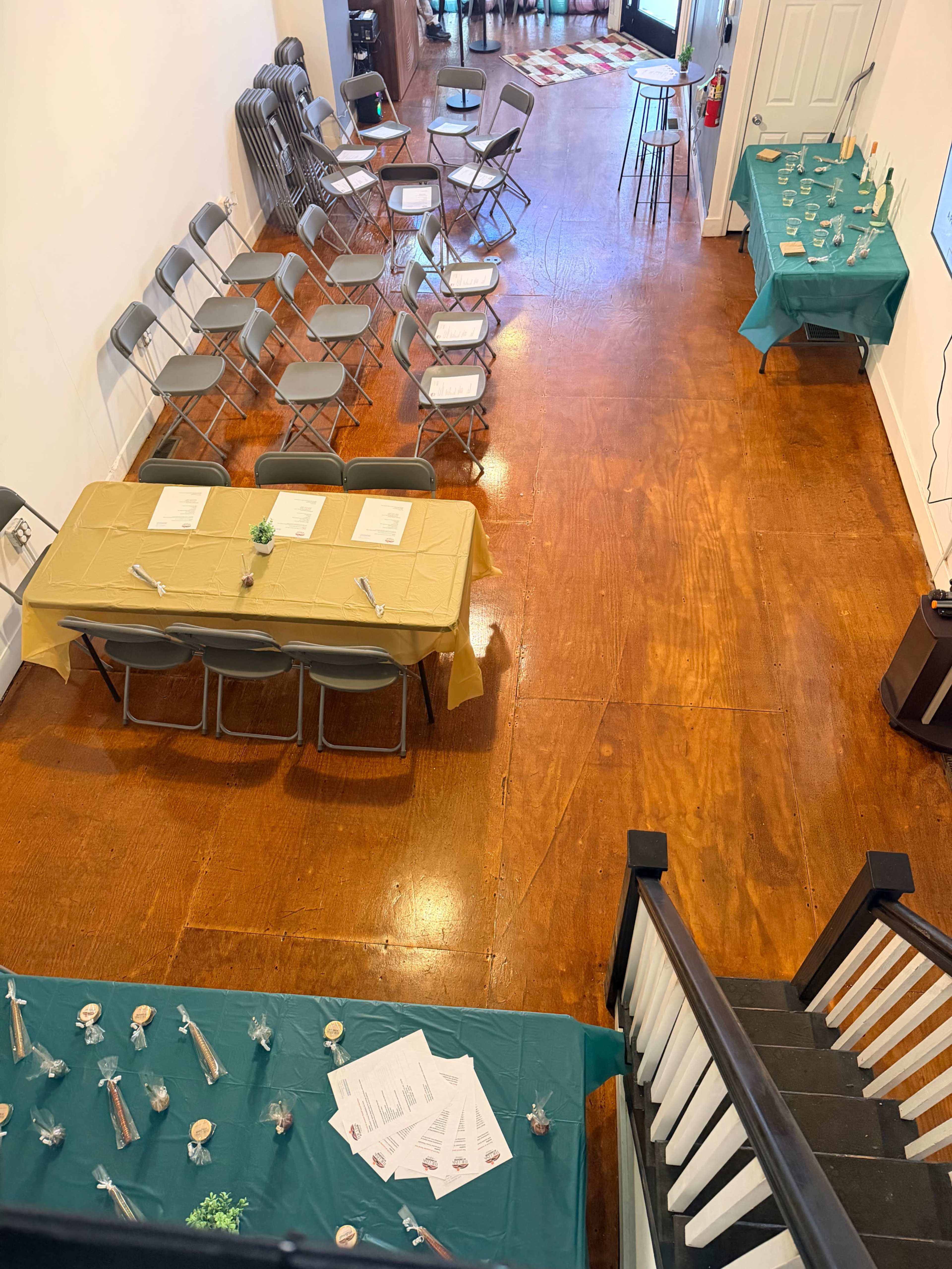 The image shows a room set up for an event, featuring rows of folding chairs, tables covered with teal and yellow cloths, and decorations arranged on the tables.